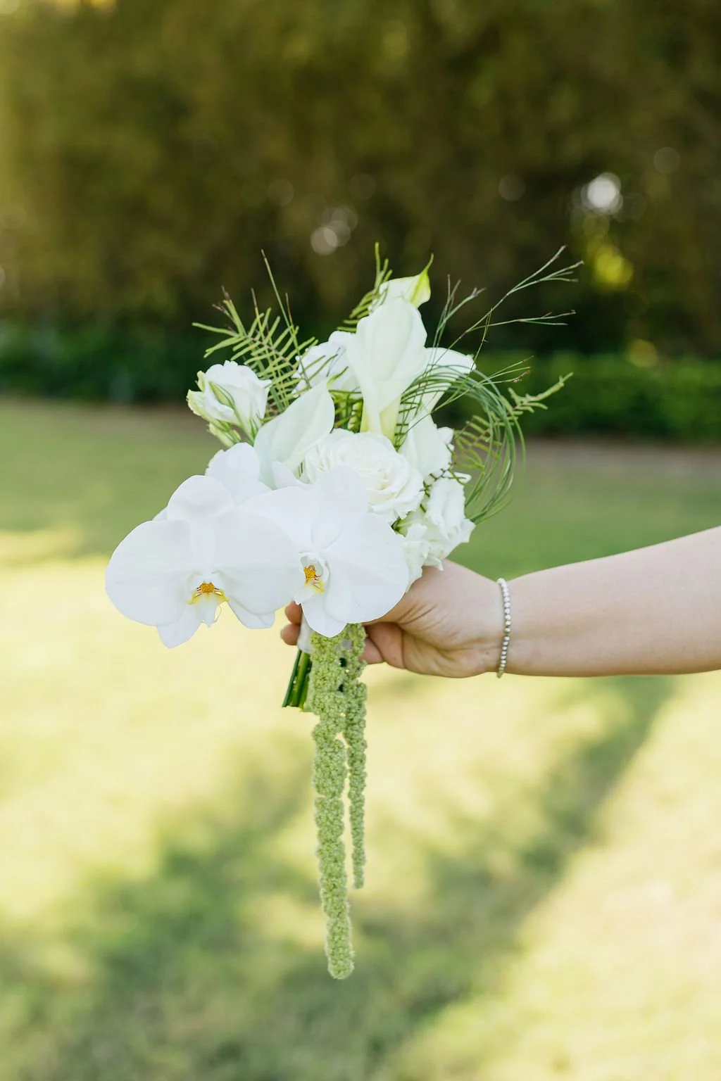 A hand holding a white floral bouquet with orchids, roses, and greenery outdoors.