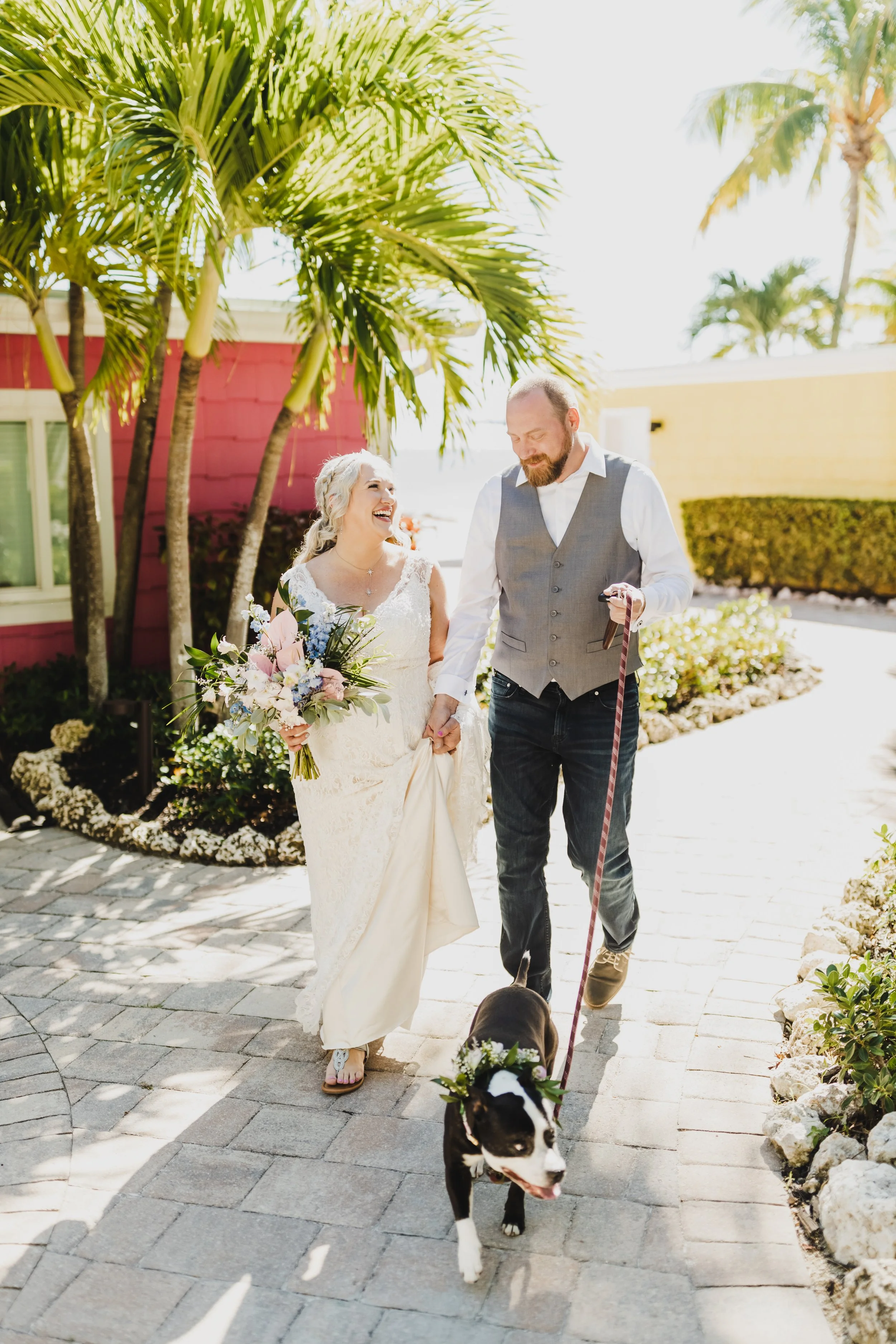 Bride and groom walking with a dog on a sunny path lined with palm trees, with the bride holding a bouquet and the dog wearing a floral wreath.