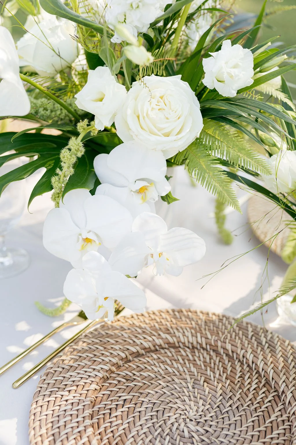A floral arrangement of white flowers including roses and orchids, with green ferns and leaves, positioned atop a white surface with a woven placemat in the foreground.