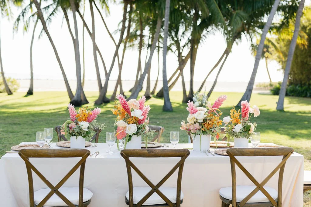 A beautifully set outdoor dining table with four floral centerpieces, glassware, and cutlery, located in a lush green park with palm trees and a water body in the background.