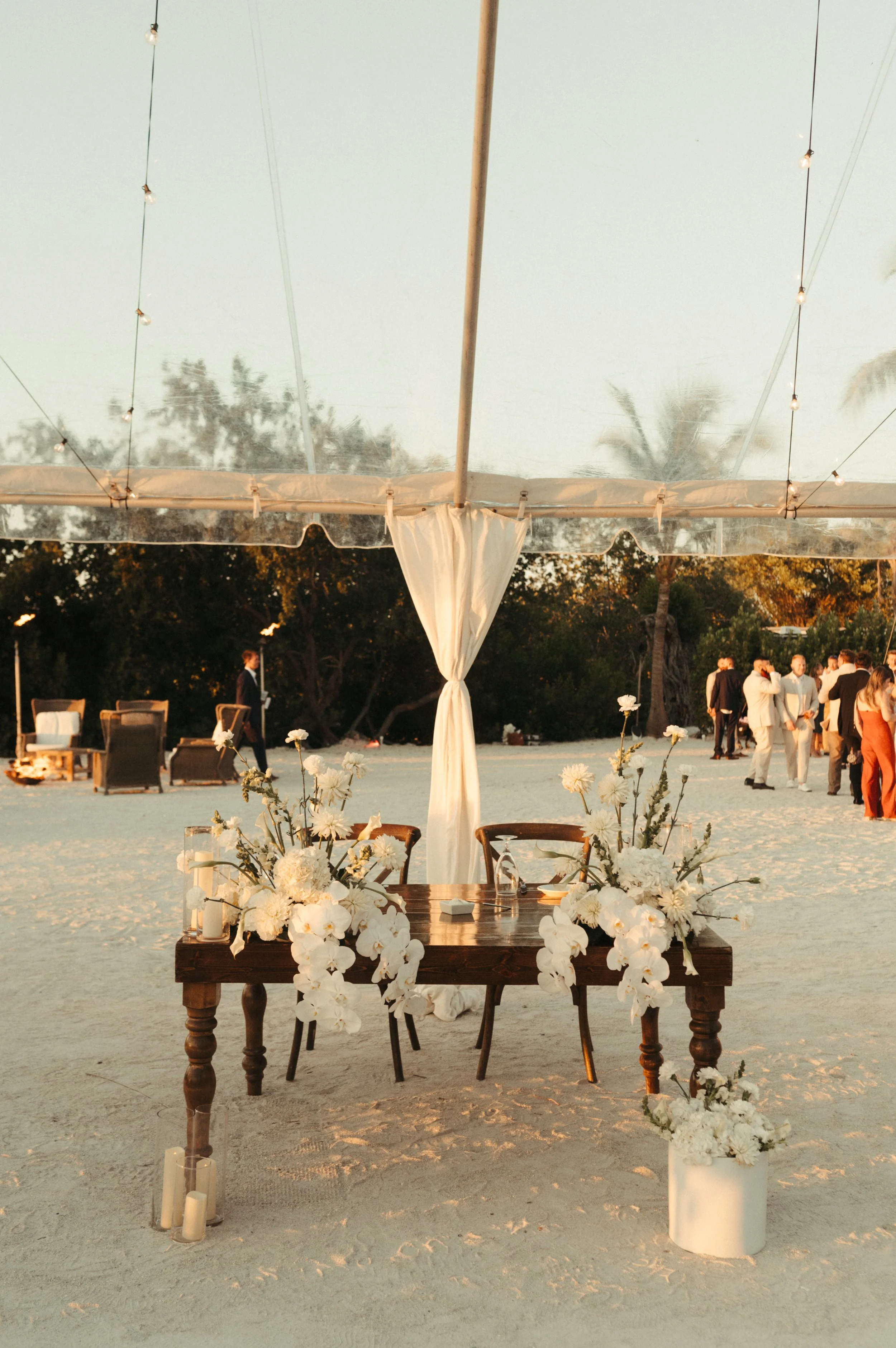 Beach wedding reception setup under a clear tent with a wooden table adorned with white flowers and candles, with people mingling in the background.