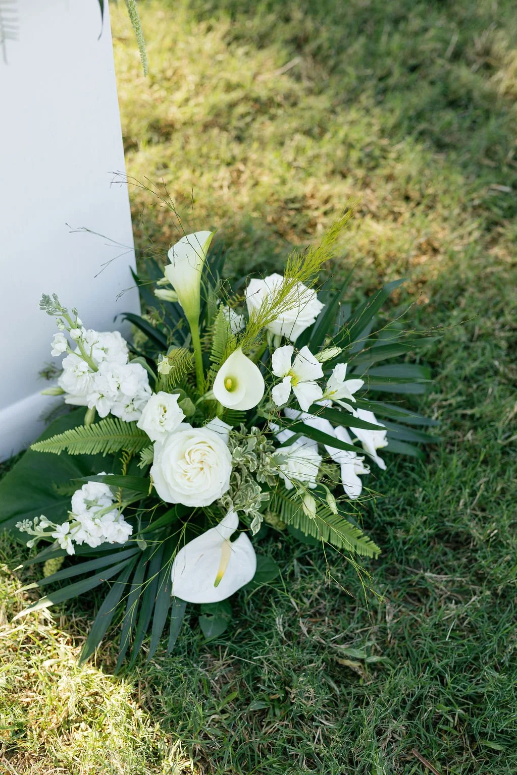 White flower arrangement with calla lilies, roses, orchids, and greenery outdoors on grass.