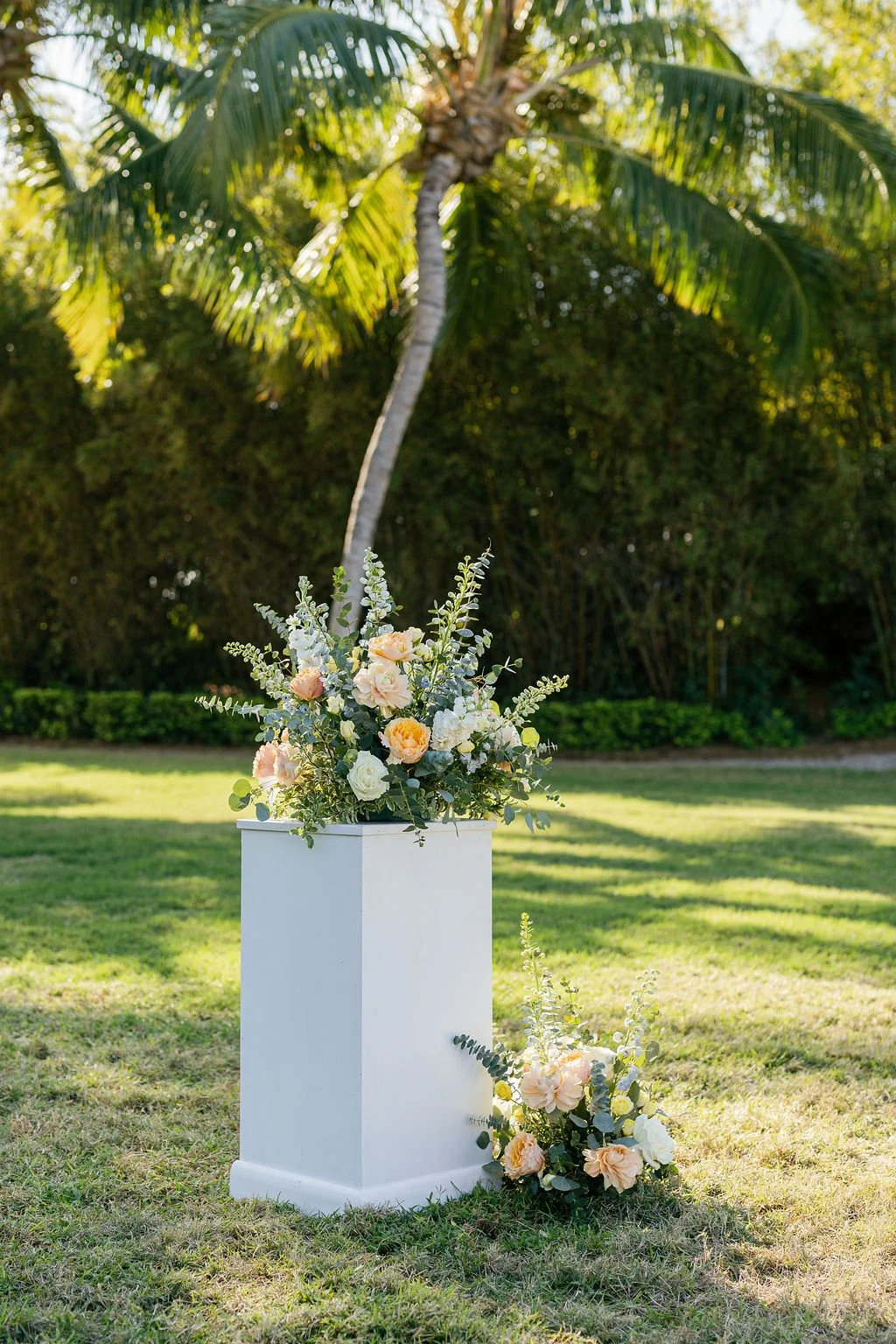 A floral arrangement with pastel-colored roses and greenery on a white pedestal outdoors, with a green lawn, bushes, and palm trees in the background.