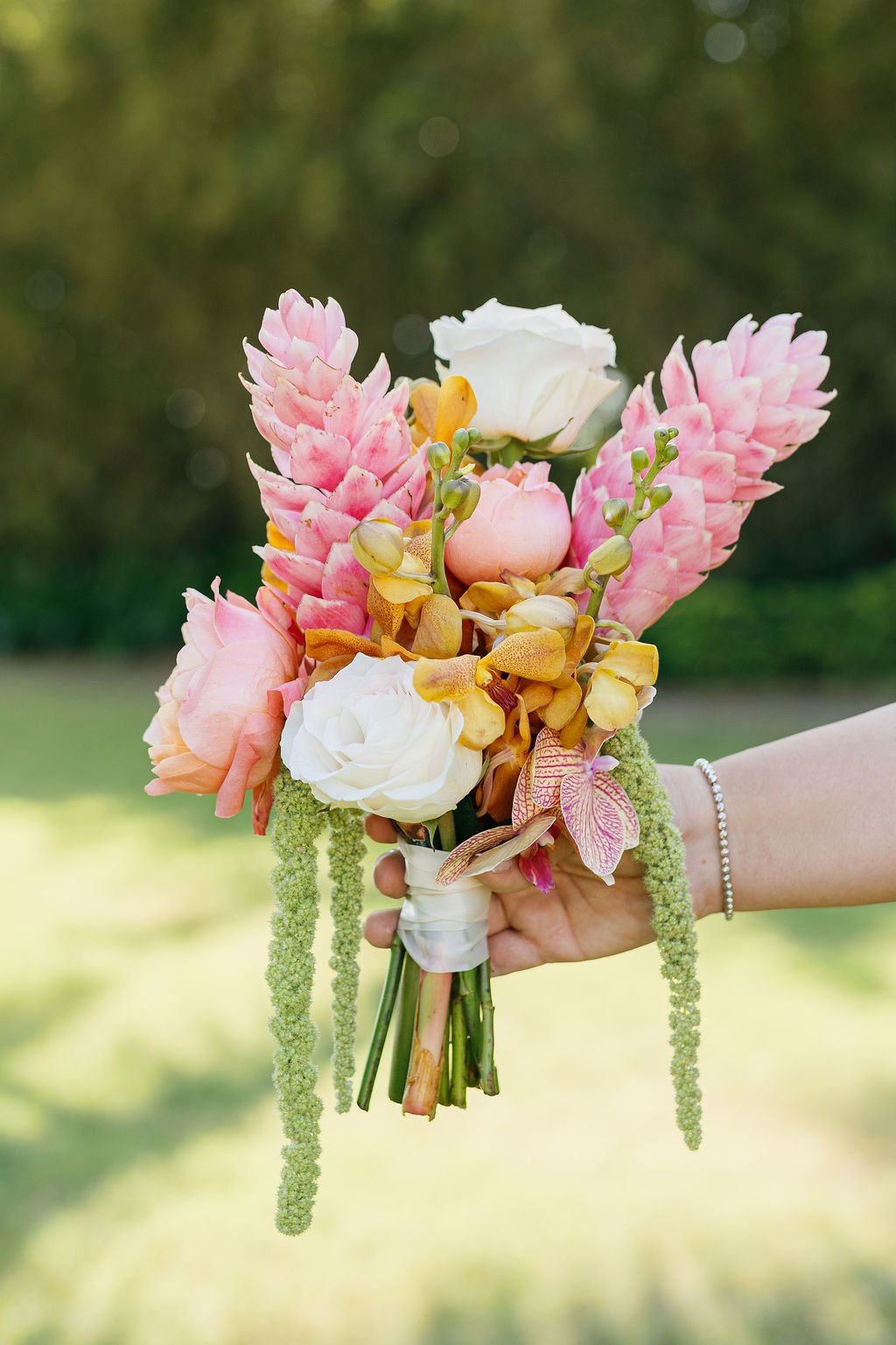A hand holding a bouquet of pink, white, yellow, and peach flowers in an outdoor setting.