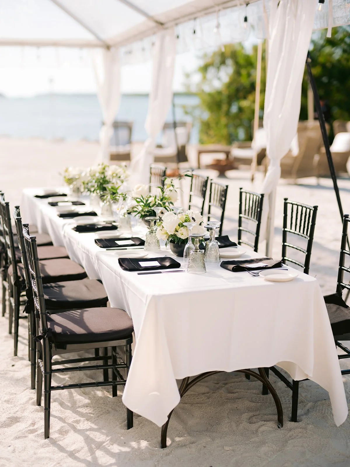 Outdoor beachside wedding table setup with white tablecloth, black chairs, and floral centerpieces under a transparent tent.