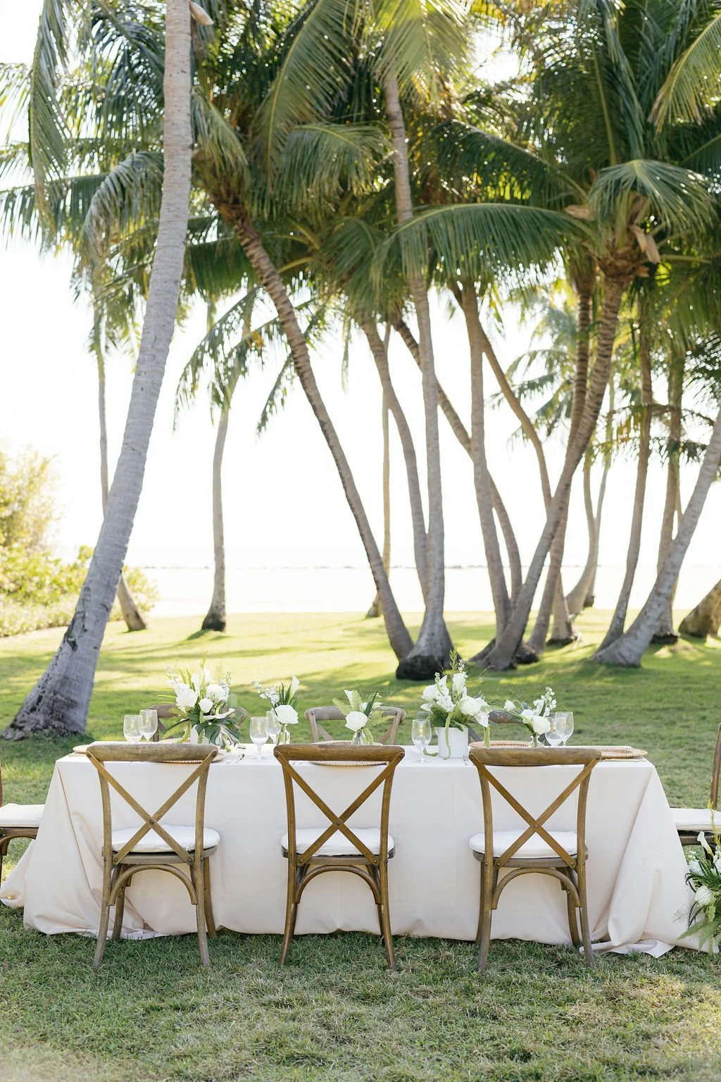 Table set for outdoor event with floral arrangements on a grassy lawn under tall palm trees.