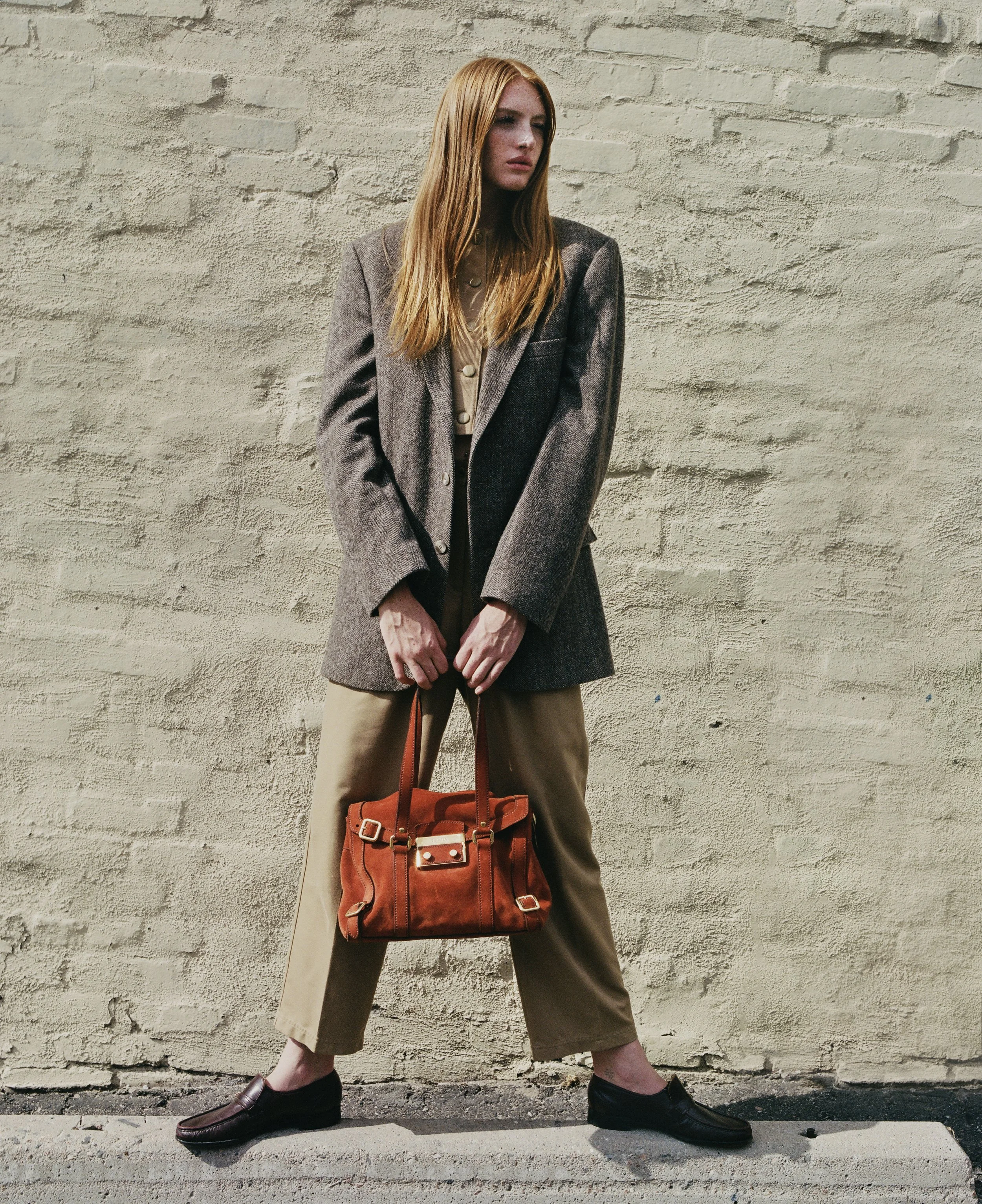 A woman with long red hair standing against a textured beige wall. She wears a brown blazer, beige pants, and black loafers. She holds an orange handbag and looks to the side with a neutral expression.