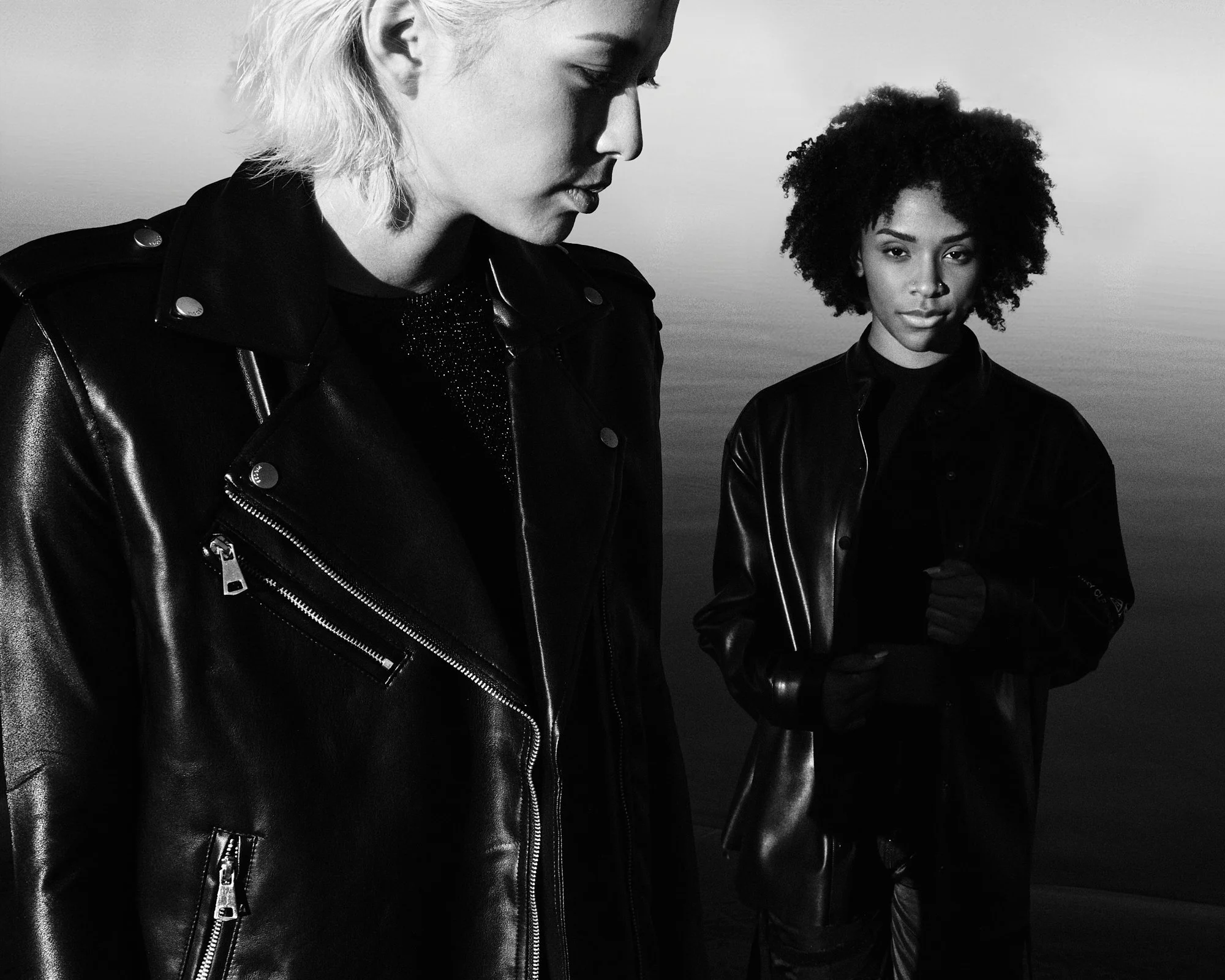 Black and white photo of two women wearing leather jackets, standing outdoors near water, one with short blonde hair looking down and the other with curly hair looking at the camera.