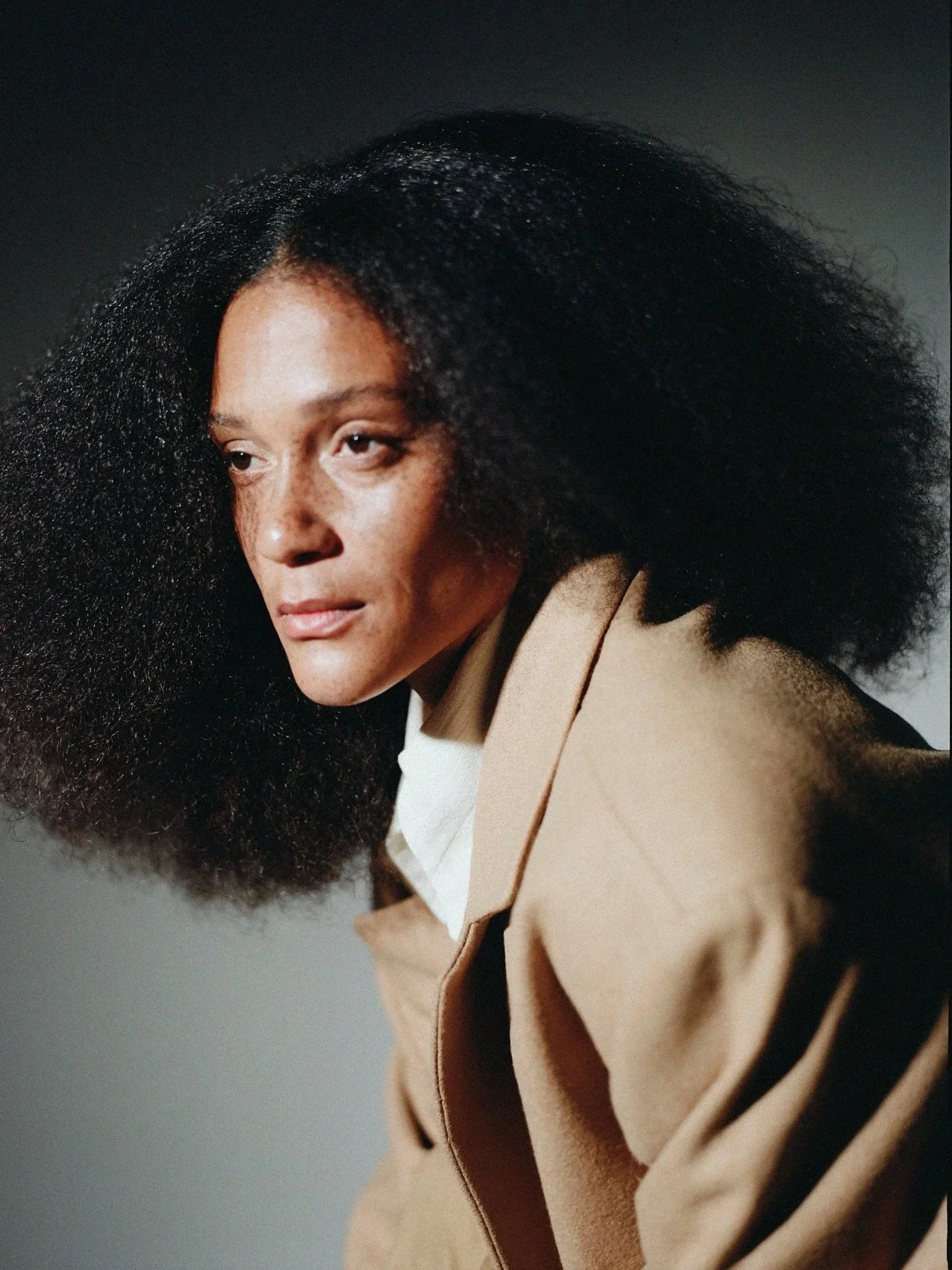 A woman with dark curly hair wearing a beige coat with black stripes on the sleeves.