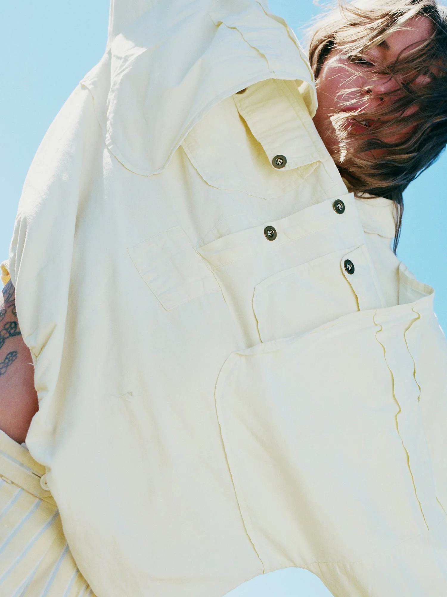 A person wearing a light yellow button-up shirt with ear-length hair, standing against a bright blue sky.