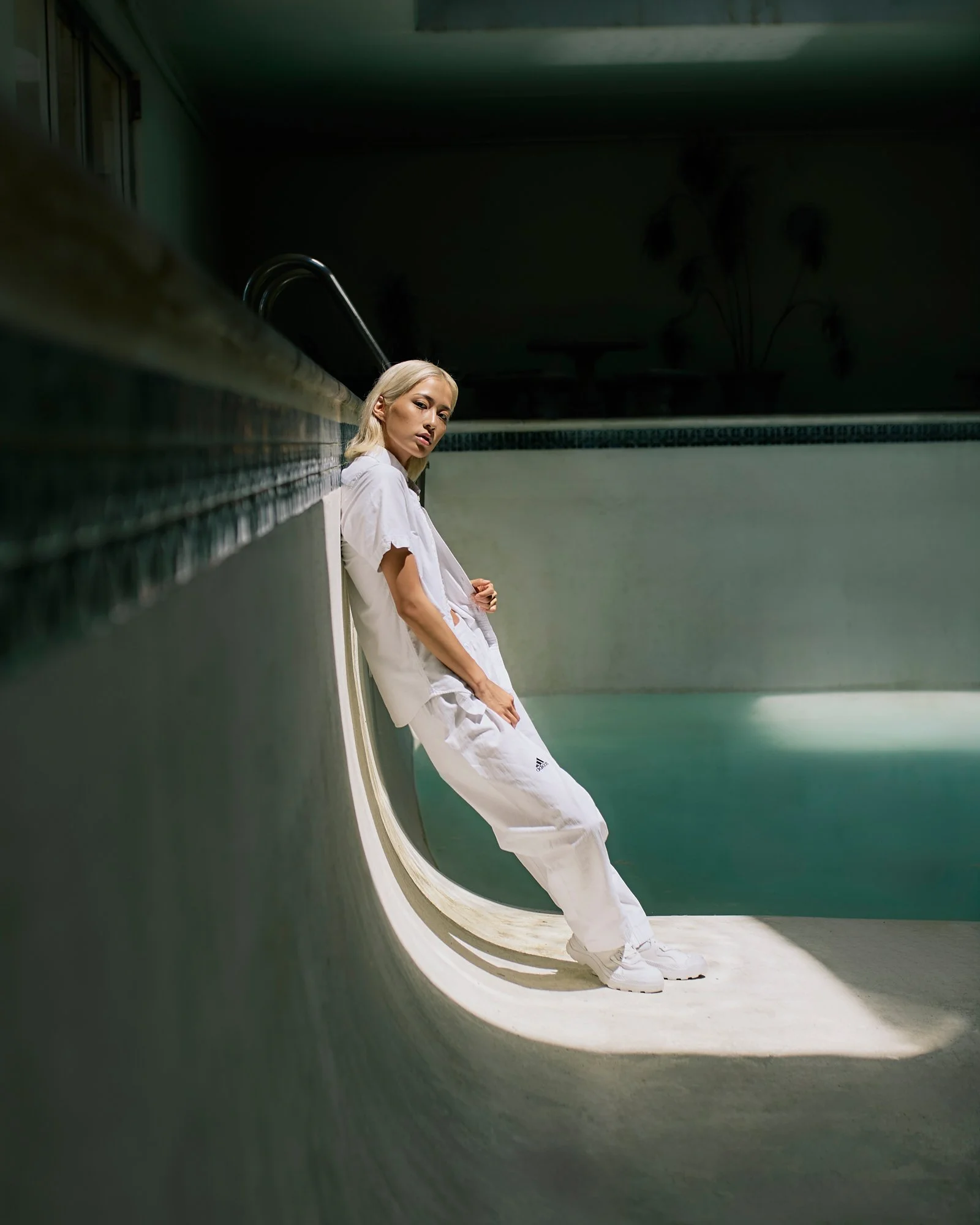 A woman with blonde hair leaning on the edge of an empty swimming pool, wearing a white outfit and sneakers, looking towards the camera.