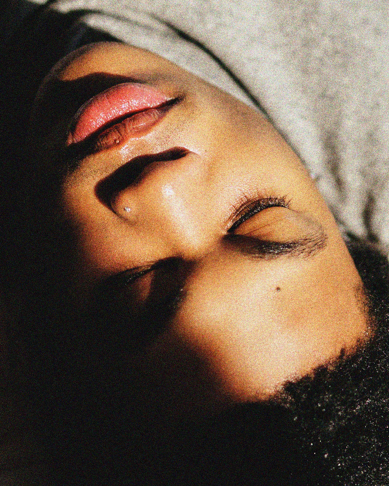 Close-up of a person's face lying with eyes closed, showing makeup and facial features, with dark hair and a light gray shirt.