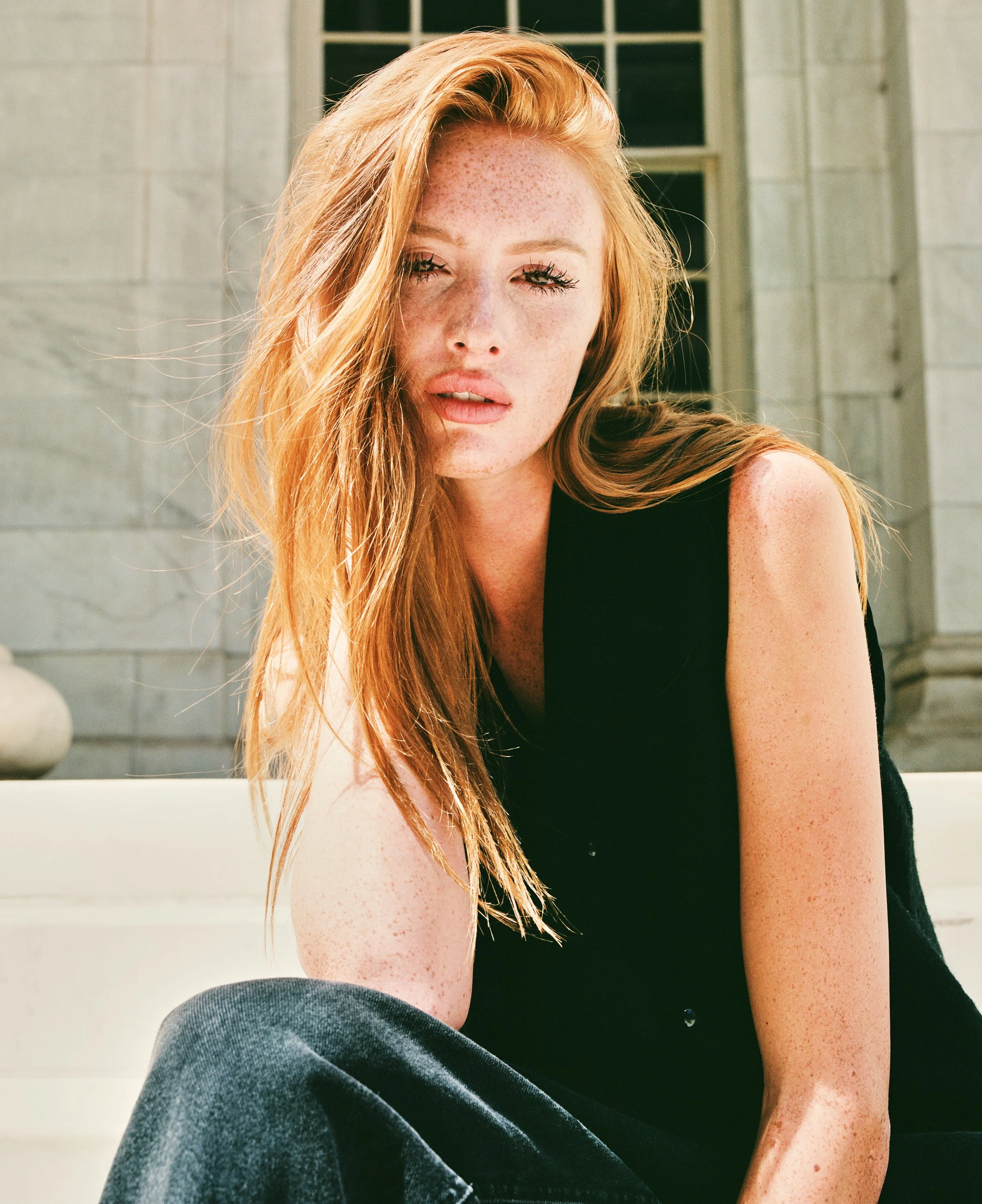 A young woman with long red hair and freckles squinting in bright sunlight, sitting outside near a building with large windows and a marble wall.