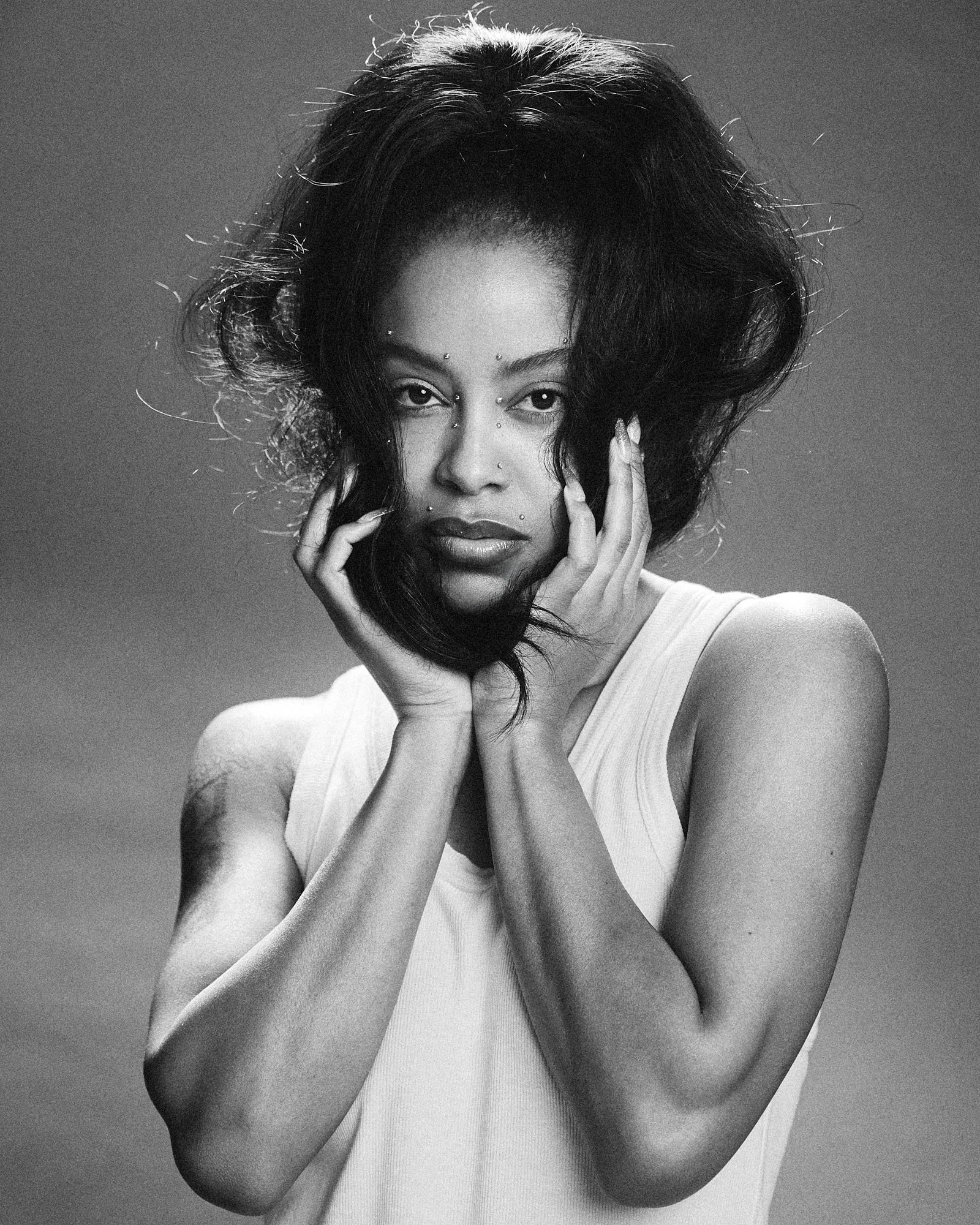 A black and white portrait of a woman with voluminous hair and facial piercings, wearing a sleeveless top, looking directly at the camera with her hands framing her face.