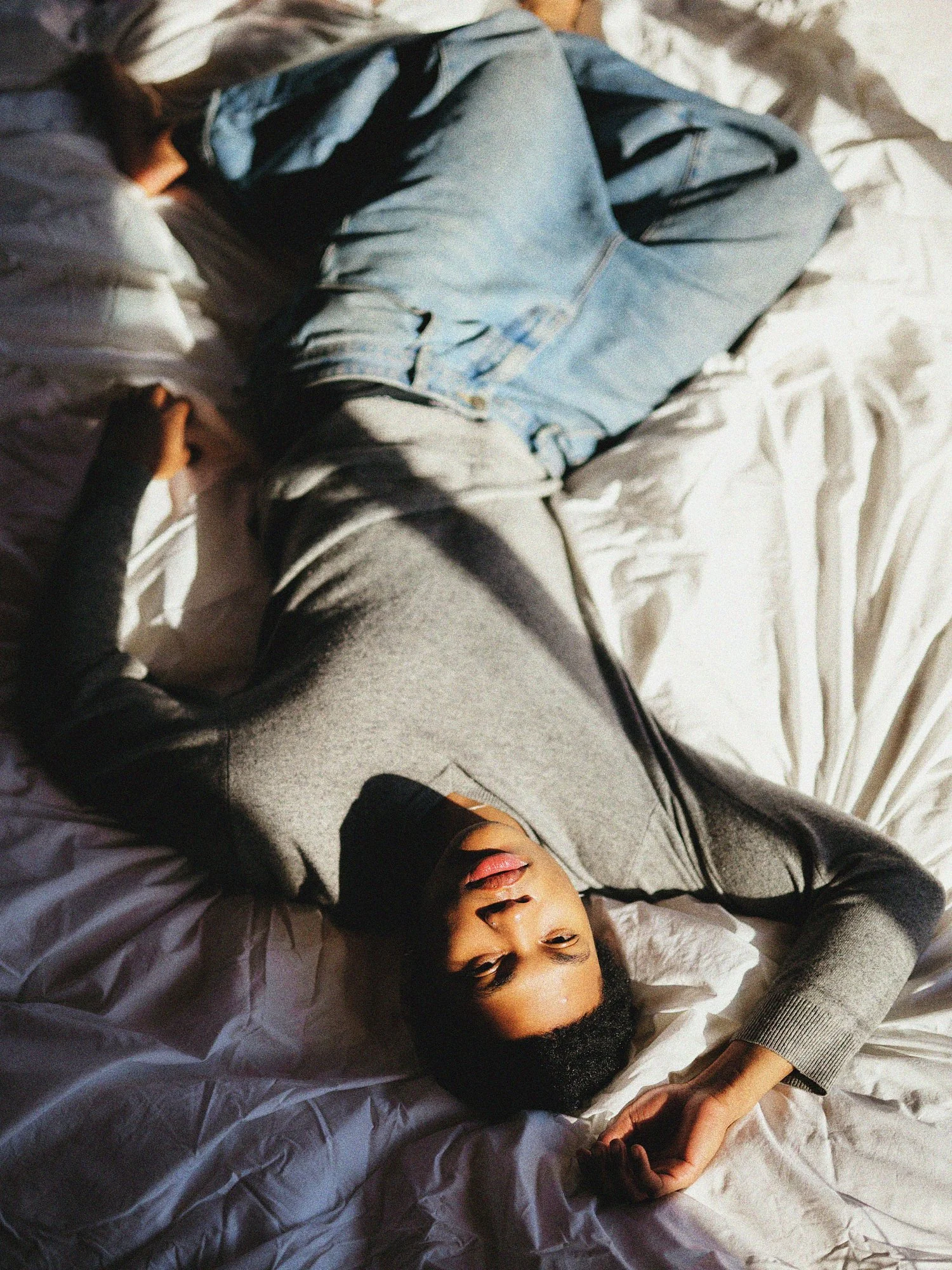 A woman with dark hair and light skin lying on a bed, wearing a gray sweater, and looking at the camera with her hand near her face.