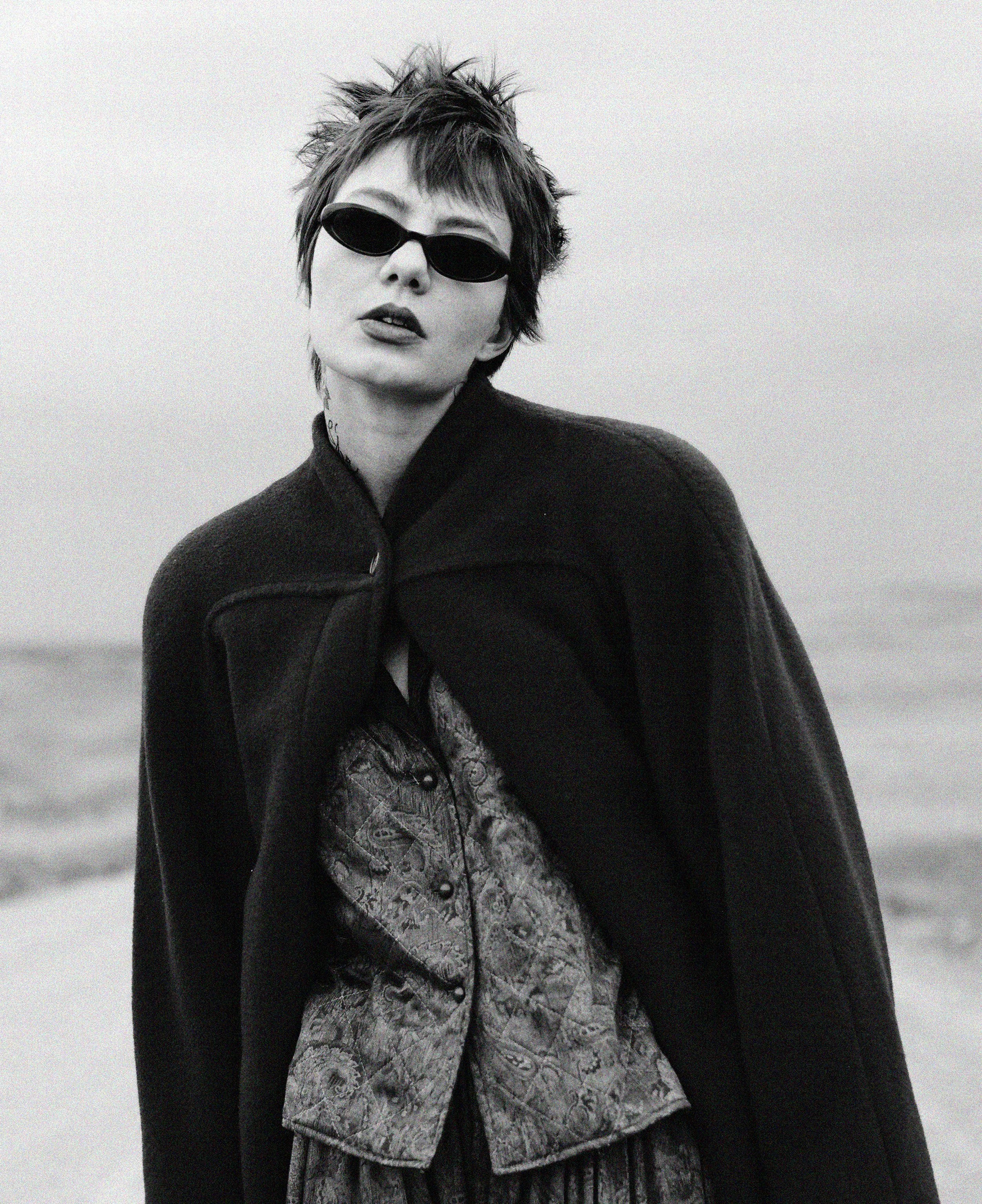 A woman with short, spiky hair wearing black sunglasses and a dark coat, standing outdoors with a blurred background.