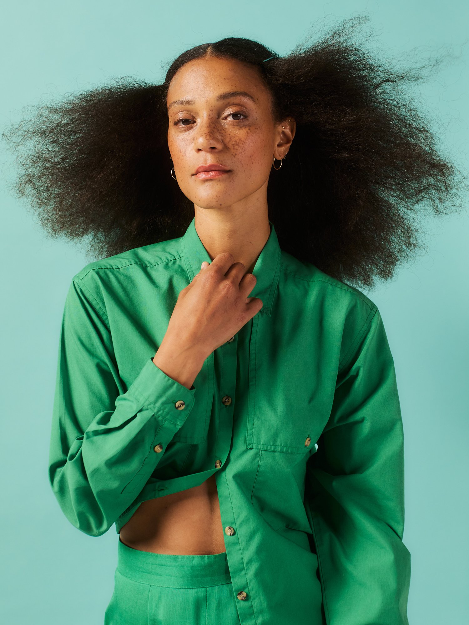 A woman with medium skin, large curly hair, and freckles wears a green shirt and matching green pants, standing against a mint green background.