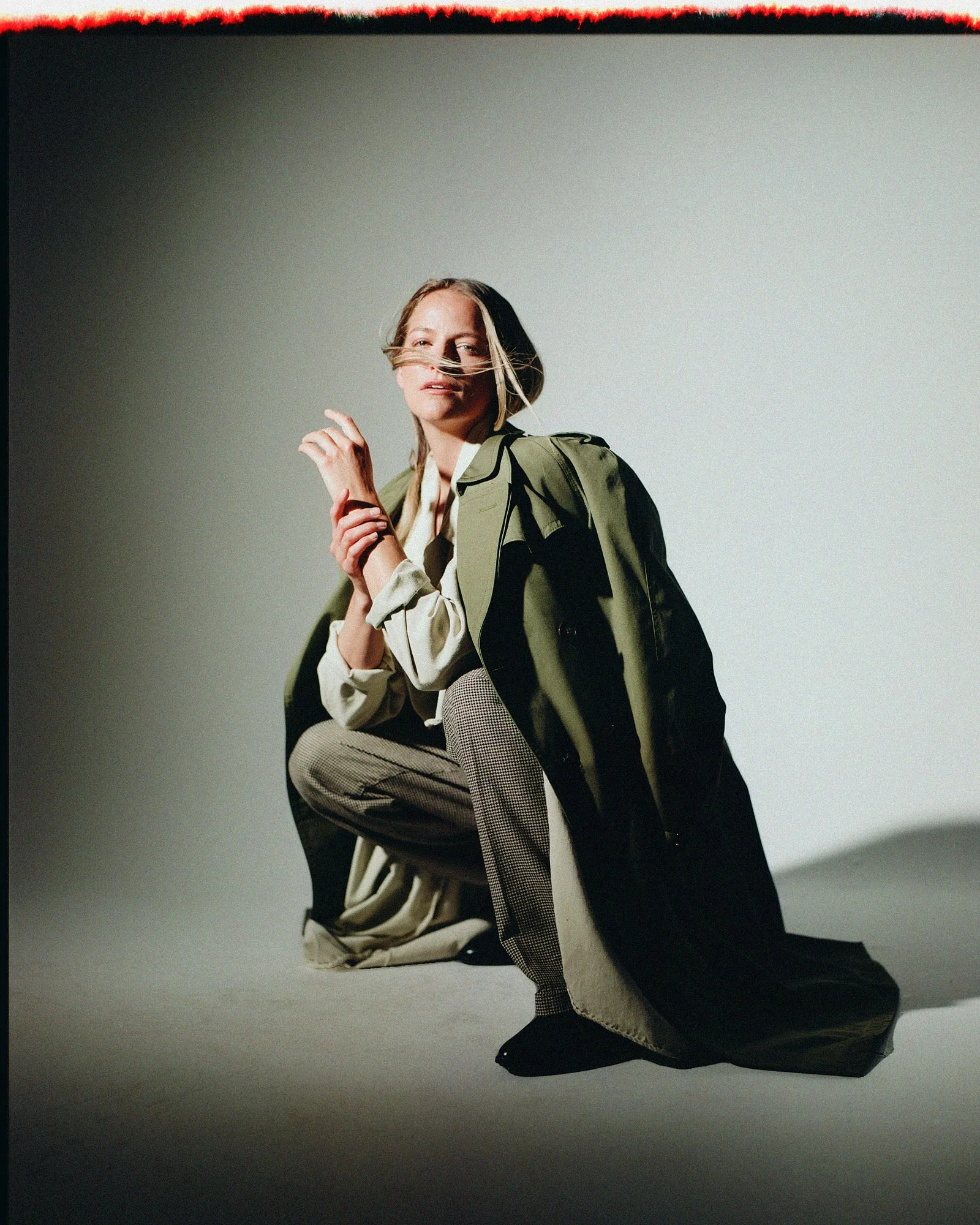 A woman with blonde hair, dressed in a beige shirt, pants, and black shoes, is kneeling with a gray and black coat draped over her shoulders and a striped bag on her lap, against a plain white background.