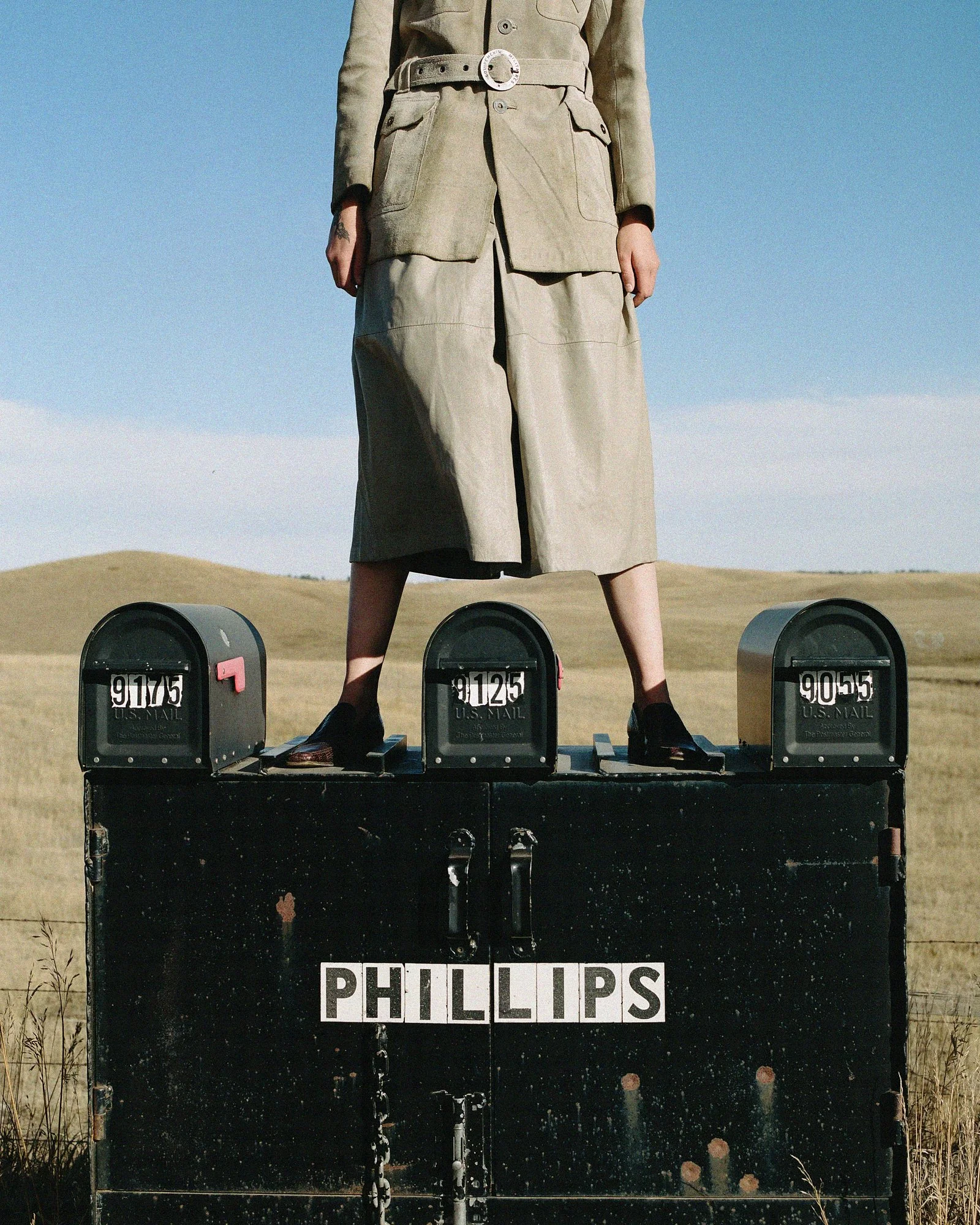 A person's lower body standing on a vintage black mailbox in an open field, with three other similar mailboxes, under a clear blue sky, with a sign reading 'PHILLIPS' on the mailbox.