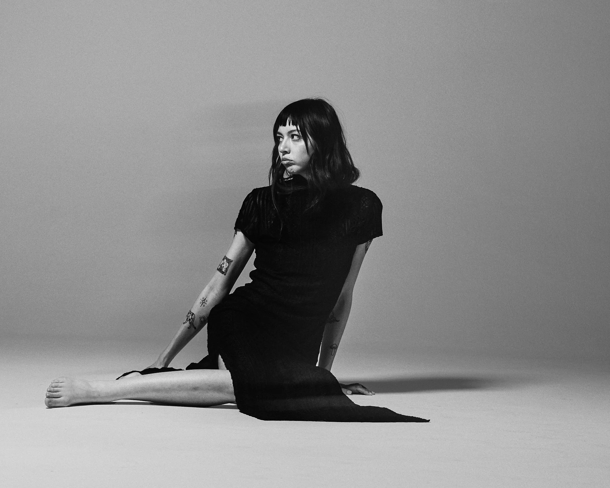 A young woman with black hair and tattoos on her arms sitting on the floor with one leg extended and the other bent, in a black dress, against a plain background.