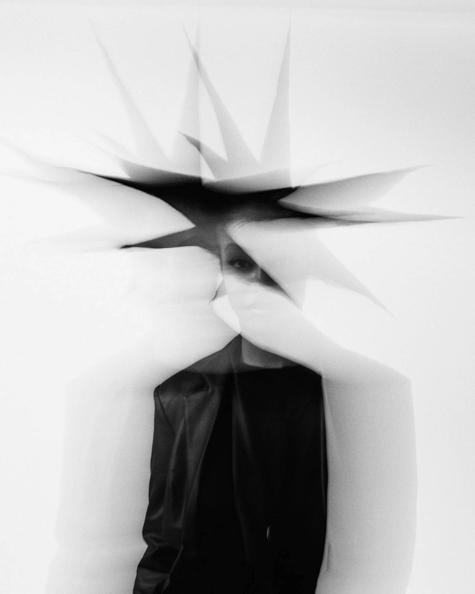 Black and white photo of a person holding a large white flower bloom close to their face, with their face partially obscured and visible only near the bottom of the frame, with a plain light background.