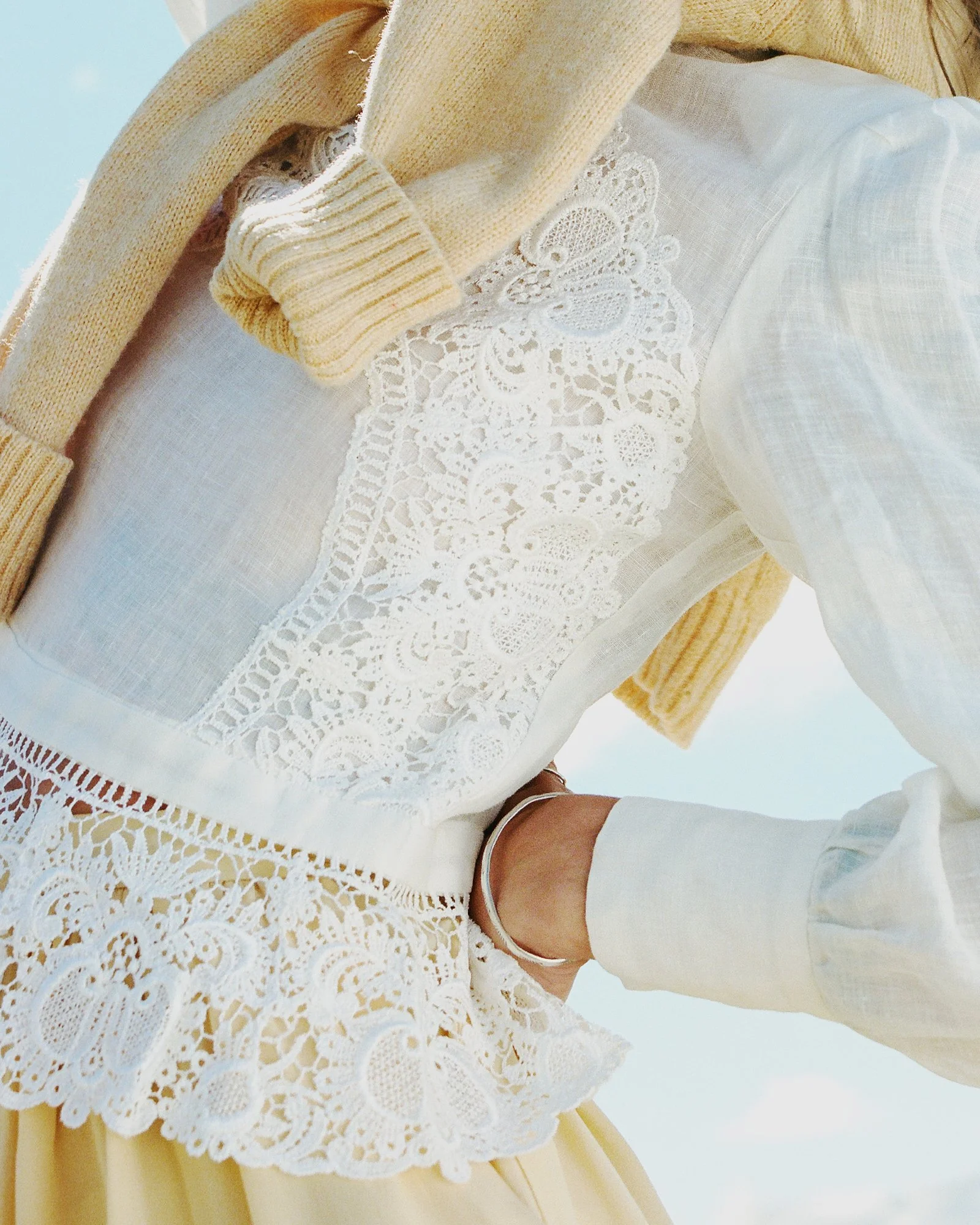 Close-up of a person wearing a yellow cardigan, a white lace top, white pants, and a silver ankle bracelet, sitting at a table with a lace tablecloth.