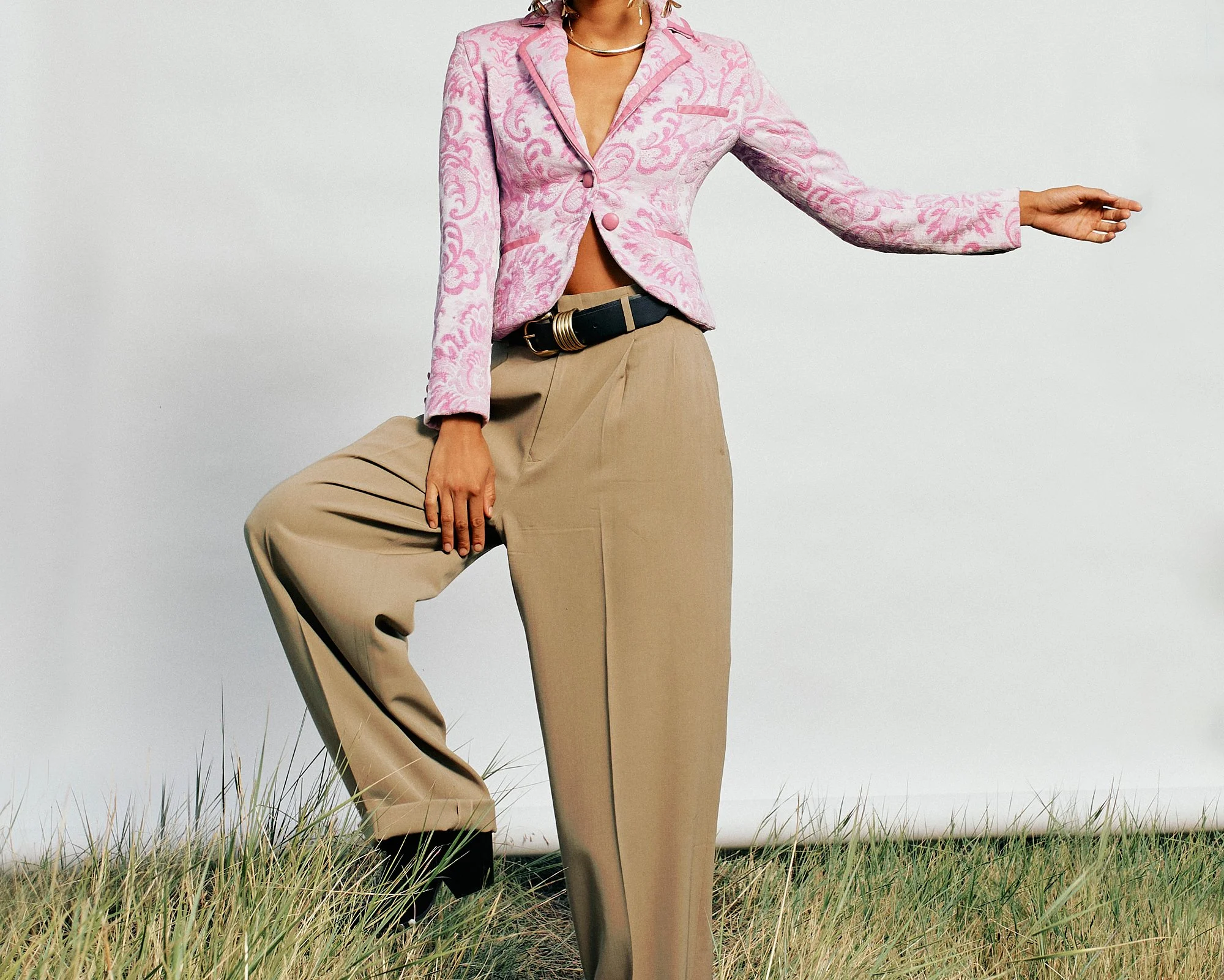 Person wearing a pink patterned blazer, beige trousers, and jewelry standing outdoors in a grassy area.