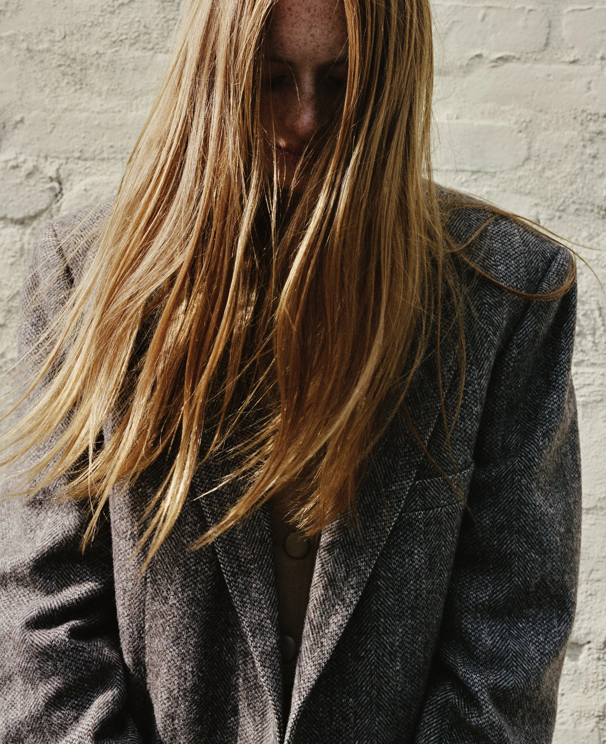 Close-up of a woman with long red hair and freckles, wearing a houndstooth blazer, standing against a white brick wall.
