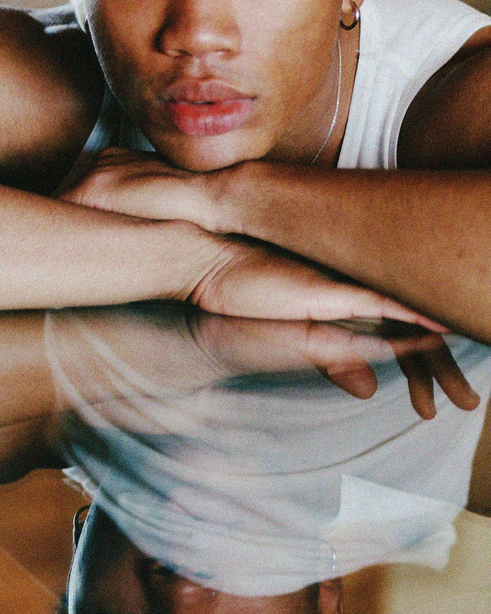 Close-up of a woman's face with lips slightly parted, wearing earrings and a thin chain necklace, leaning on her arm, with part of her white shirt and arm visible.