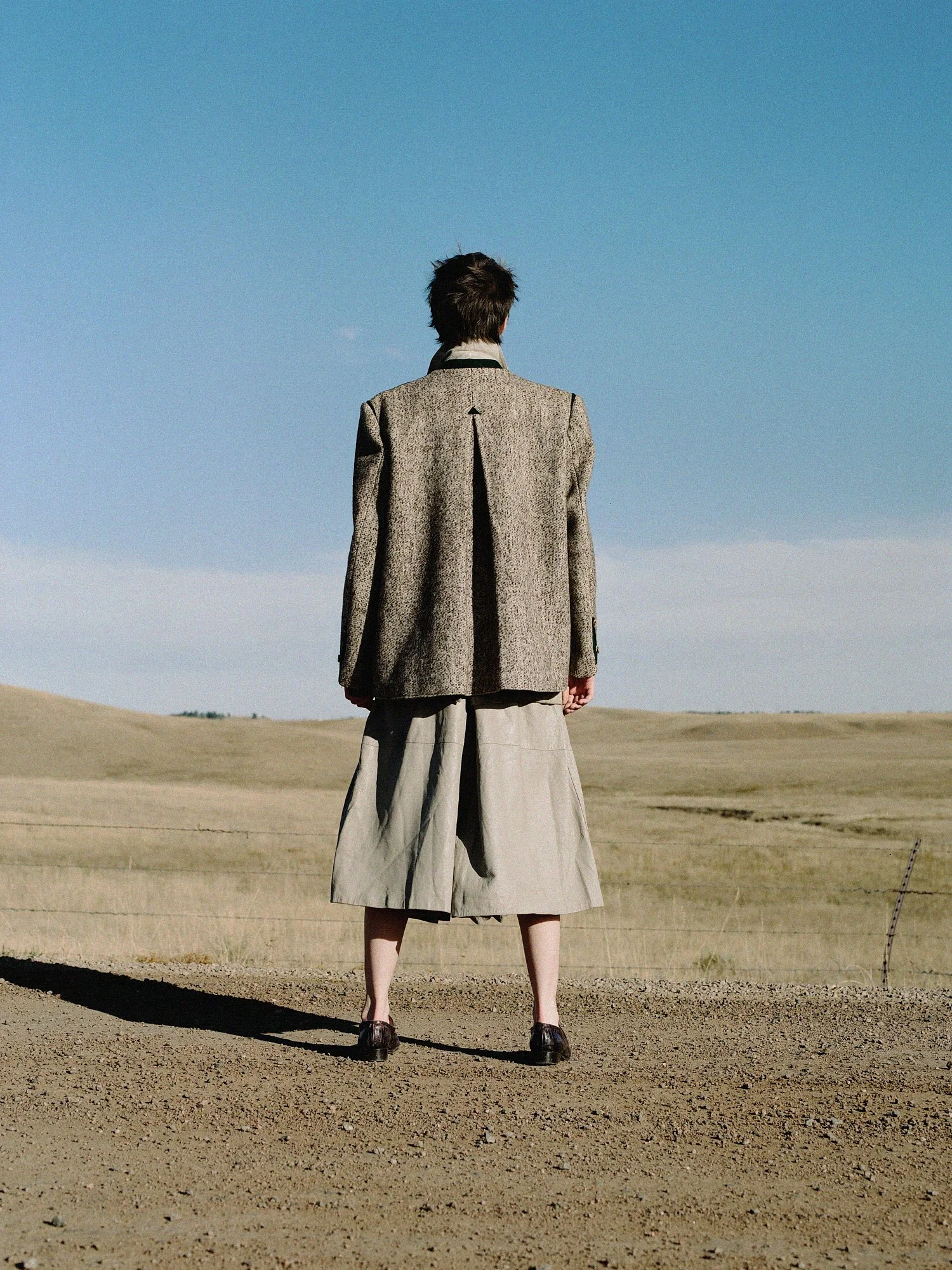 Person standing in a vast, barren desert landscape with a clear blue sky, dressed in a gray coat, wide pants, and black shoes, facing away from the camera.