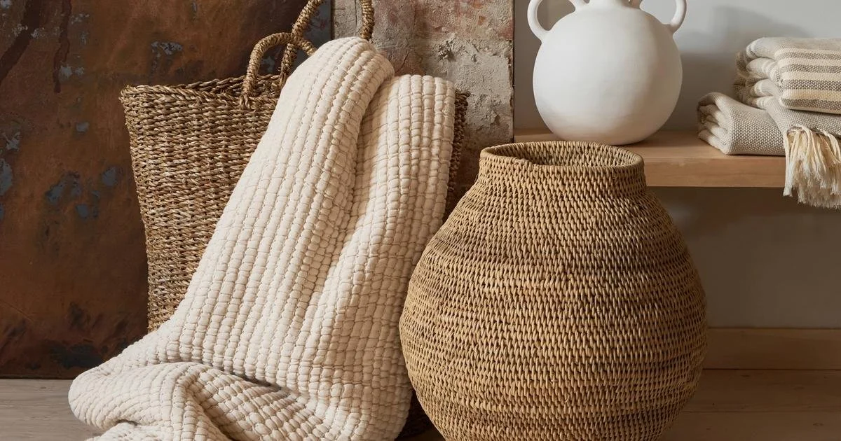 Cozy home decor arrangement with a woven basket, white ceramic vase, soft cream blanket, and folded throw pillows.