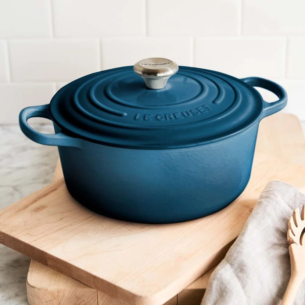 Blue Le Creuset Dutch oven with lid on wooden cutting board, next to a cloth napkin and wooden spoon.