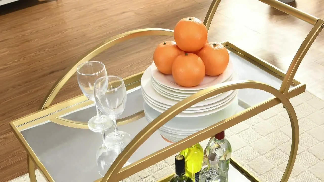 A gold-framed bar cart with stacked white plates, several oranges on top, and empty wine glasses on a glass surface, placed on a wooden floor with a beige rug underneath.