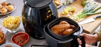 Air fryer on a kitchen counter with cooked chicken inside, surrounded by various ingredients and small dishes.