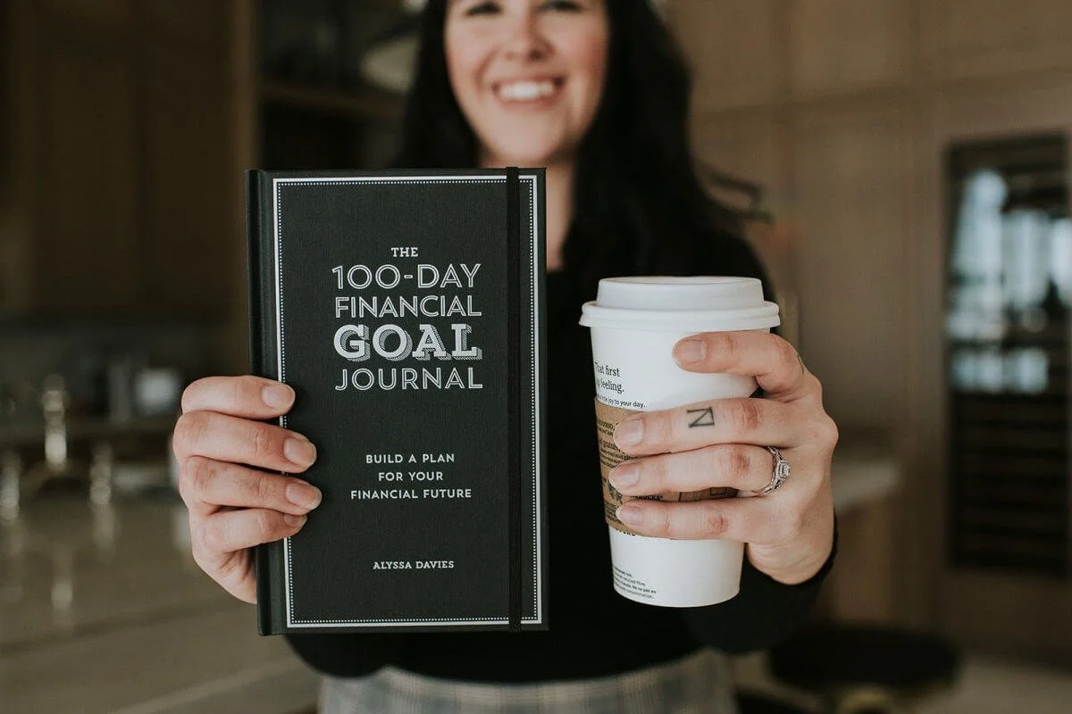Person holding 'The 100-Day Financial Goal Journal' and a coffee cup.
