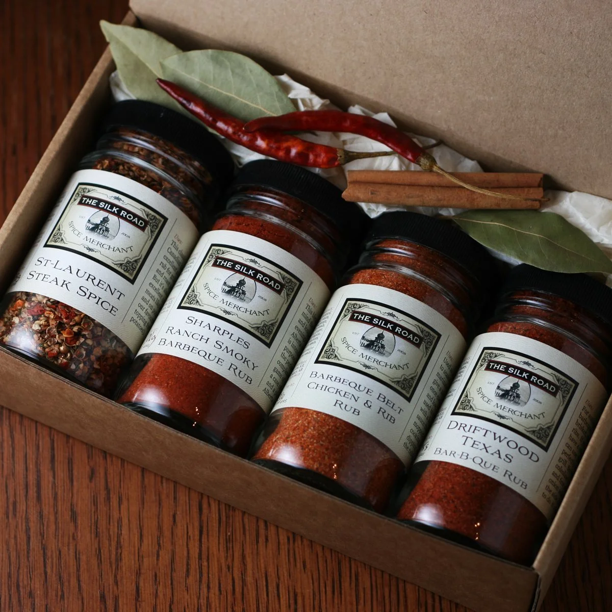 Four jars of barbecue spices labeled "The Silk Road" with varieties including St-Laurent Steak Spice, Sharples Ranch Smoky Barbecue Rub, Barbeque Belly Chicken & Rib Rub, and Driftwood Texas BBQ Rub, placed in a box with dry bay leaves, red chili peppers, and cinnamon sticks.