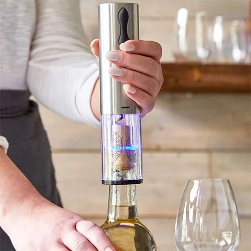 A person using an electric wine opener on a bottle of white wine, with two wine glasses nearby.