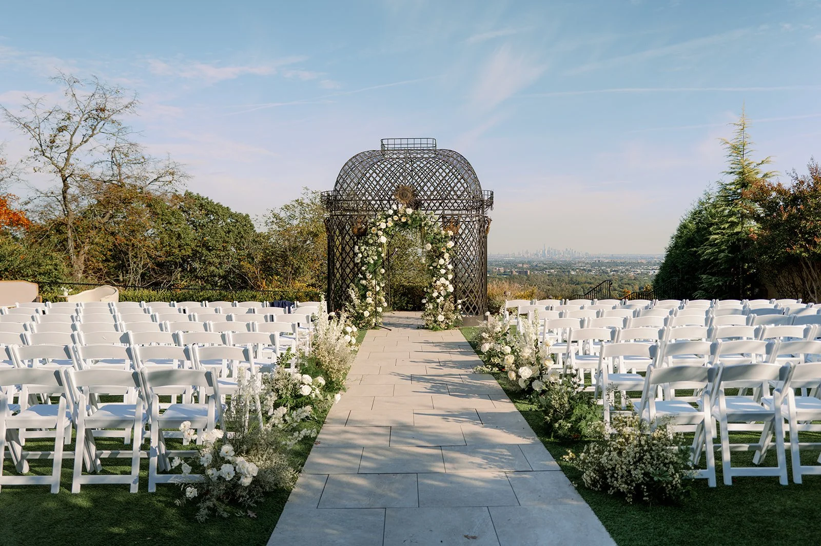 Outdoor wedding ceremony setup with white chairs, a flower-decorated arch, and a decorative gazebo on a hill with a city skyline in the distance.
