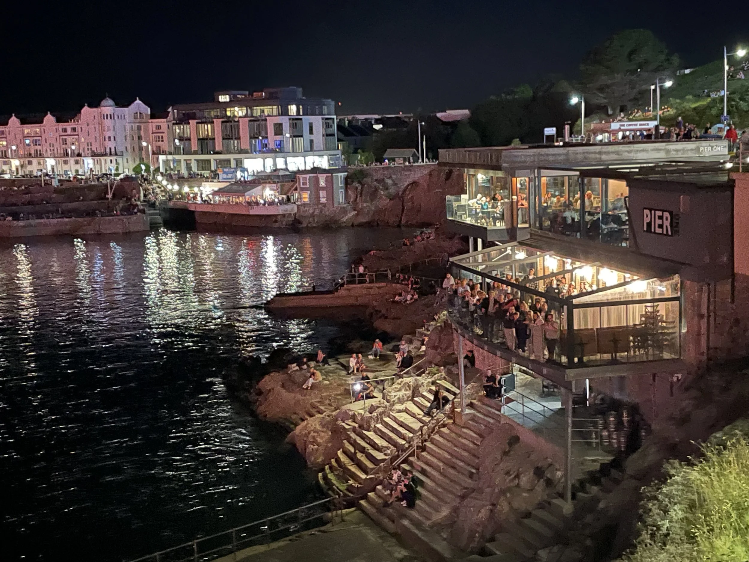 Night view of a waterfront restaurant called Pier One with large glass windows, outdoor seating, and people dining, situated on rocky stairs along the water's edge, with colorful building lights in the background.