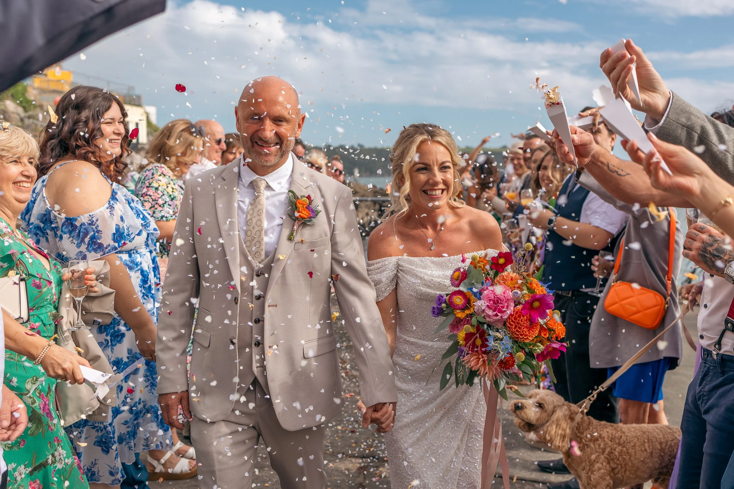A newlywed couple walks hand-in-hand through a crowd of smiling friends and family, celebrating with confetti and a dog on a leash, outdoors on a sunny day, after their wedding ceremony.
