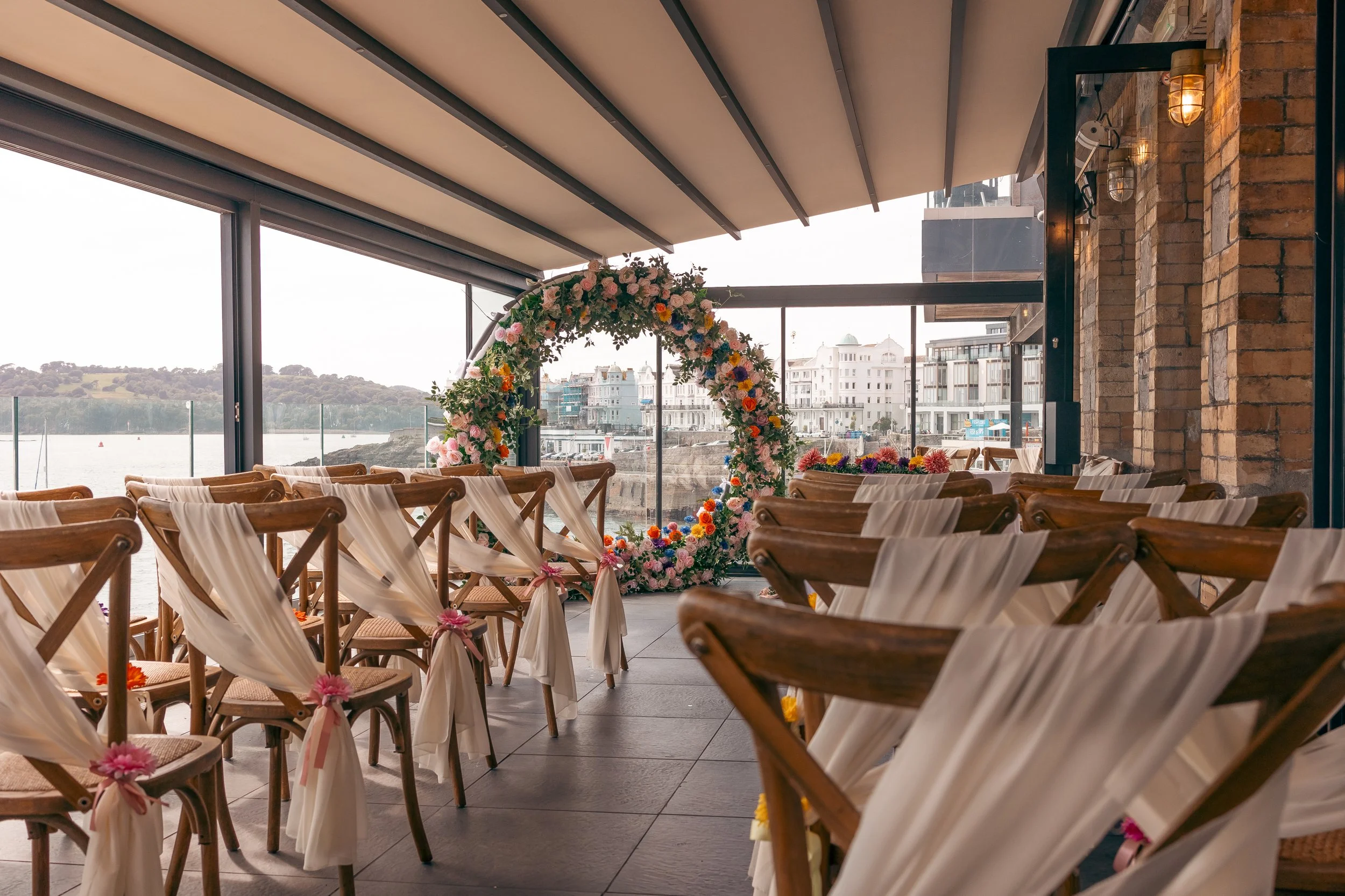 A decorated outdoor wedding ceremony setup with wooden chairs adorned with white fabric and pink bows, an arch made of colorful flowers, and a view of a waterfront with buildings in the background.