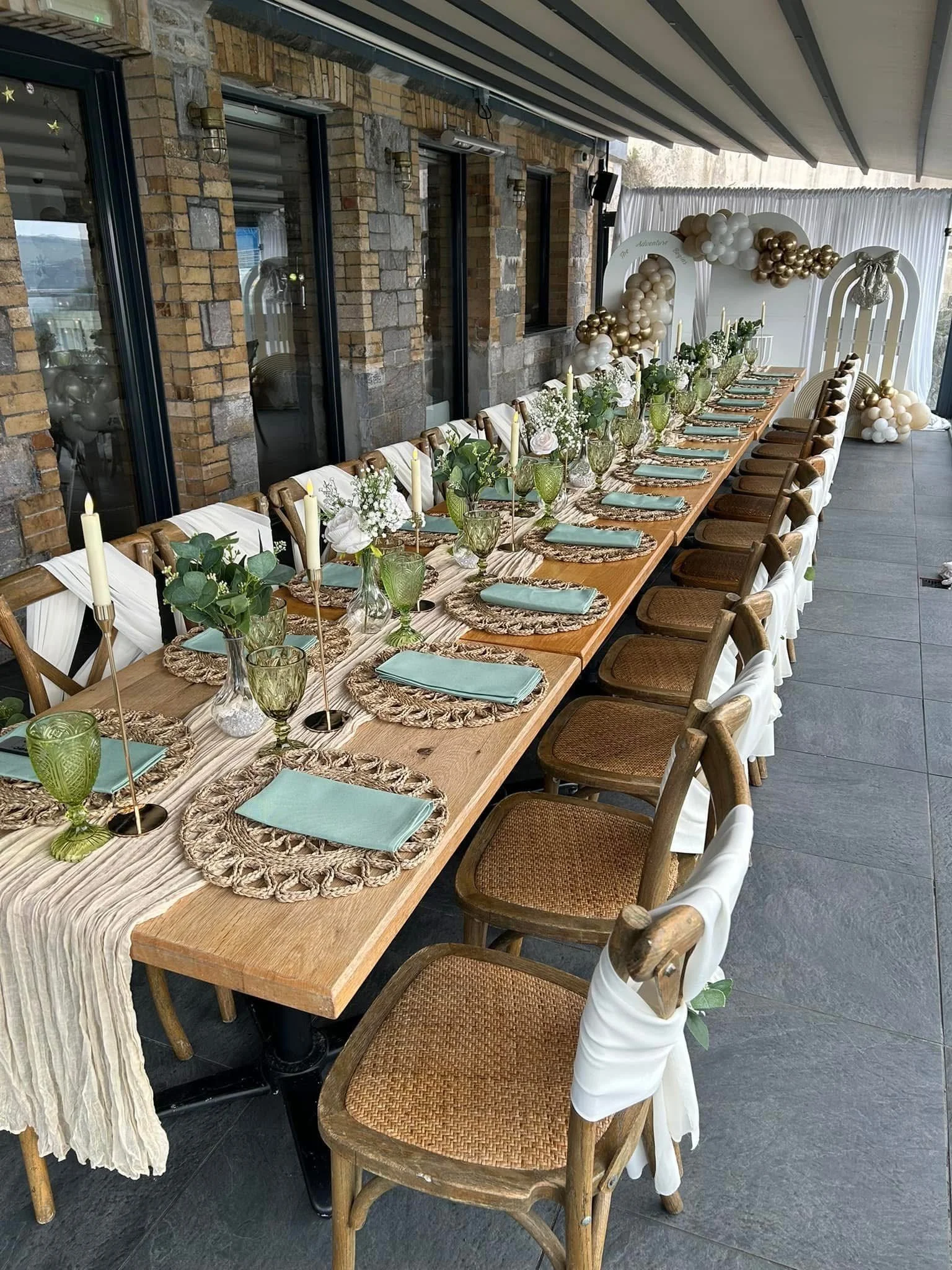 Long wooden table set for a celebration with floral arrangements, green goblets, and candles, decorated with white and gold balloon arch and backdrop.