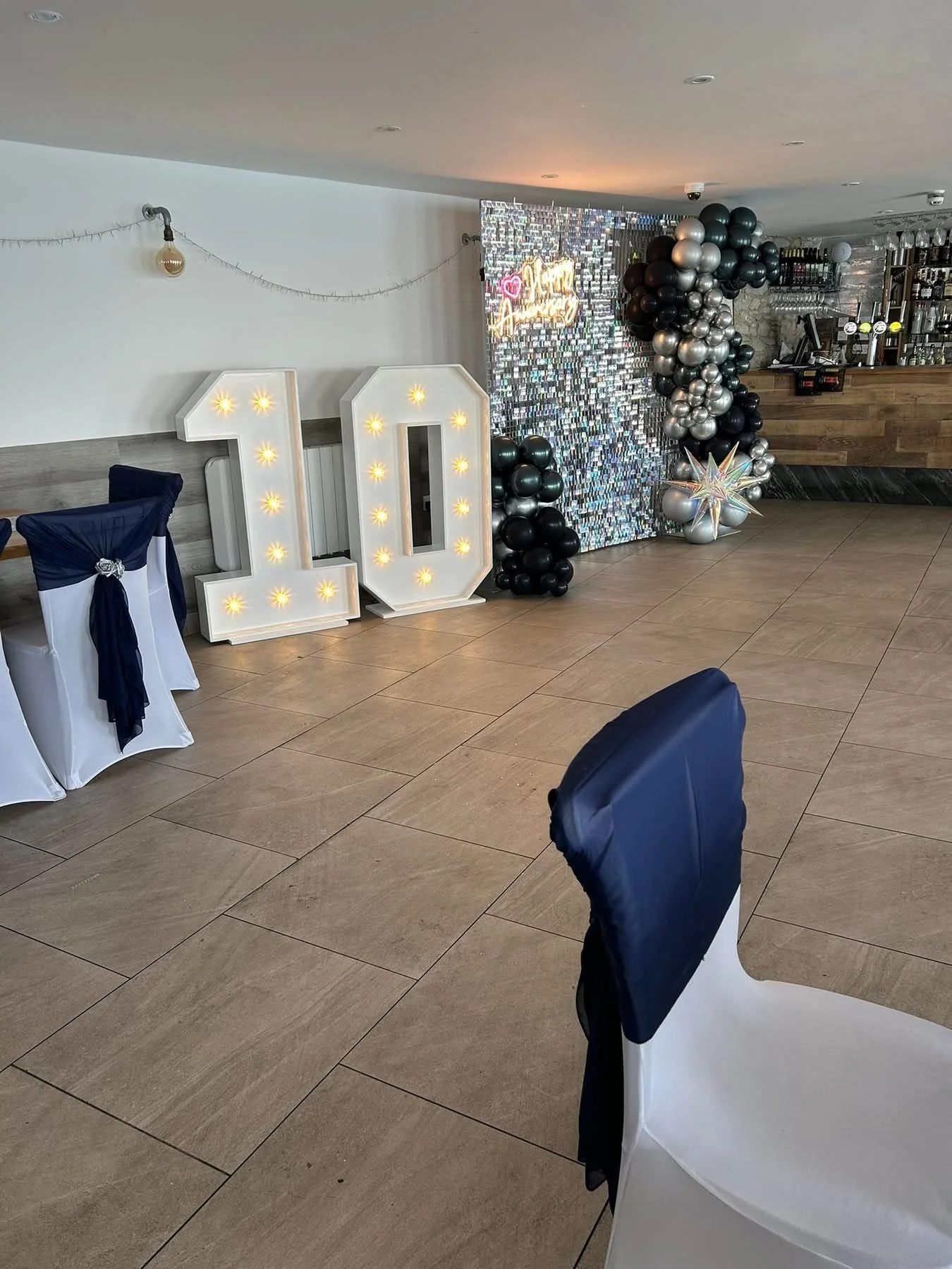 Decorations for a 10th birthday celebration with large illuminated number '10', black, silver, and gold balloons, and a reflective backdrop in a venue with chairs covered in white and navy blue cloth.