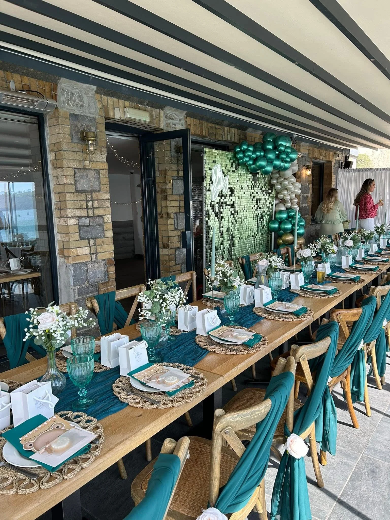 A decorated outdoor dining area with a long wooden table set for a celebration, featuring teal table runners, white flowers, gift boxes, and balloons in teal, white, and metallic gold.