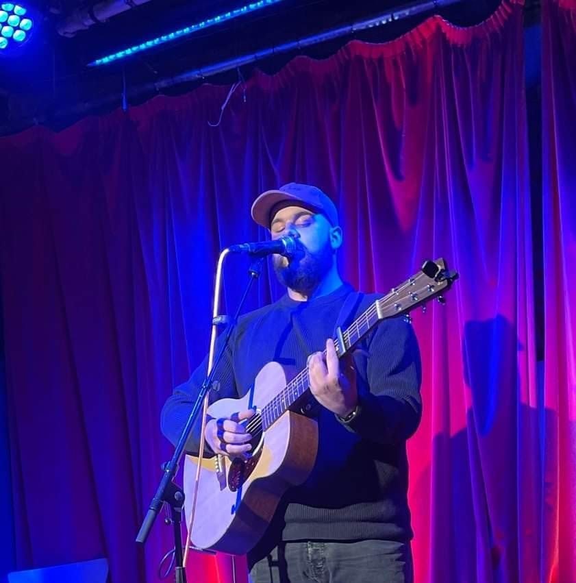 A man singing into a microphone while playing an acoustic guitar on stage with purple curtains in the background.