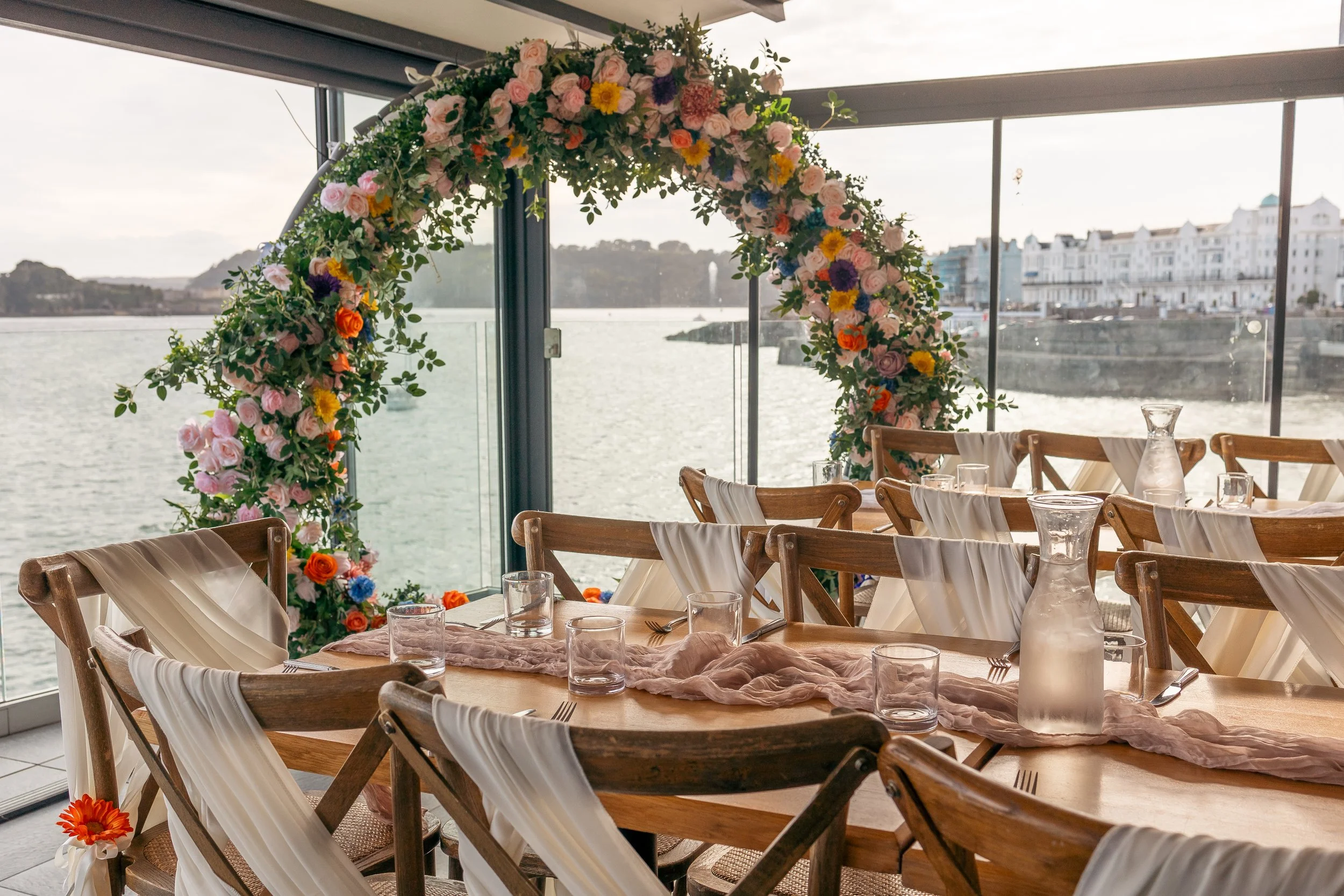 A decorated event space with a floral arch over a table near a waterfront, with wooden chairs, glassware, and a pink table runner.
