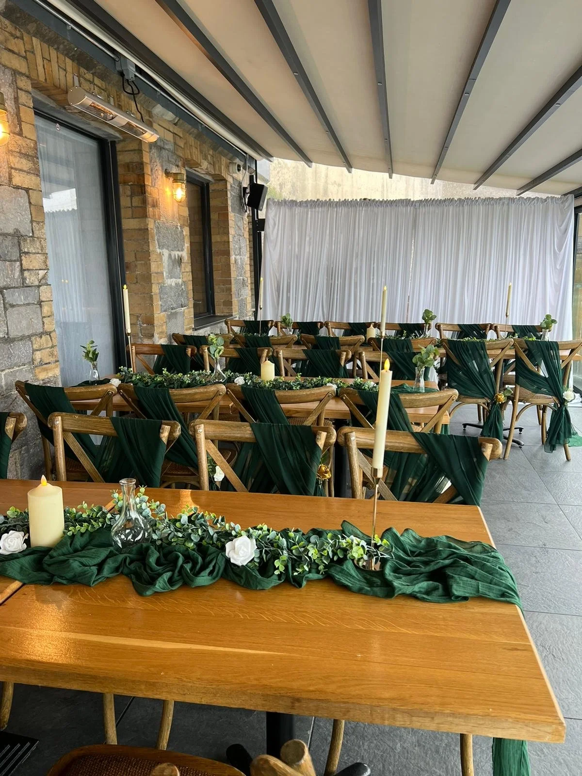 Decorated outdoor event space with wooden tables and chairs, green fabric accents, candles, and floral arrangements under a covered patio.
