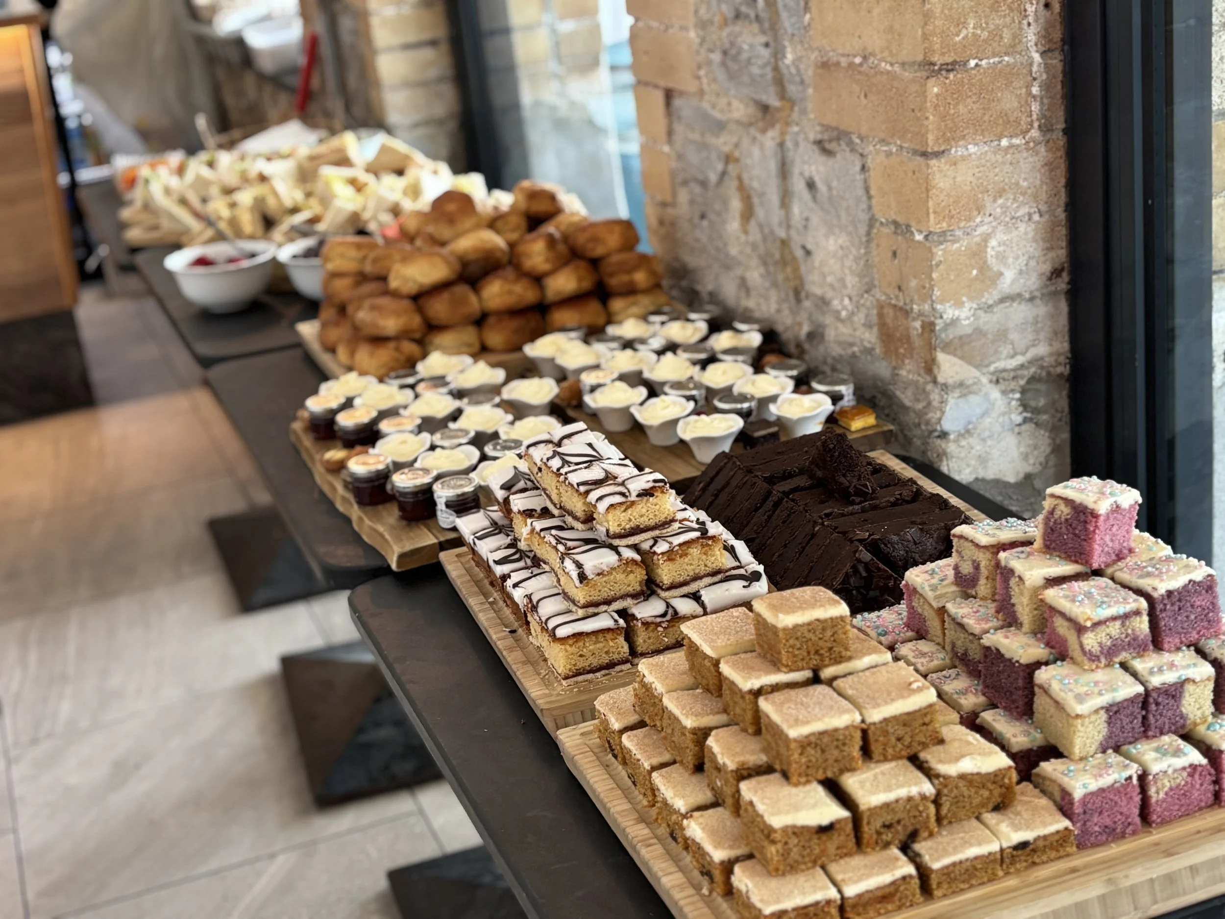 A table with an assortment of baked goods and desserts, including brownies, frosted cake squares, petit fours, mini cheesecakes, and fried food, set against a rustic brick wall and large windows.