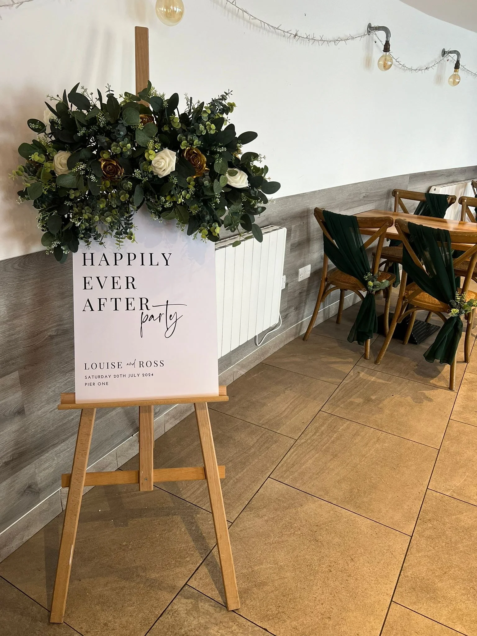 Wedding reception decoration with a floral sign on a wooden easel, and chairs decorated with green fabric and small flower arrangements, in a modern indoor setting.