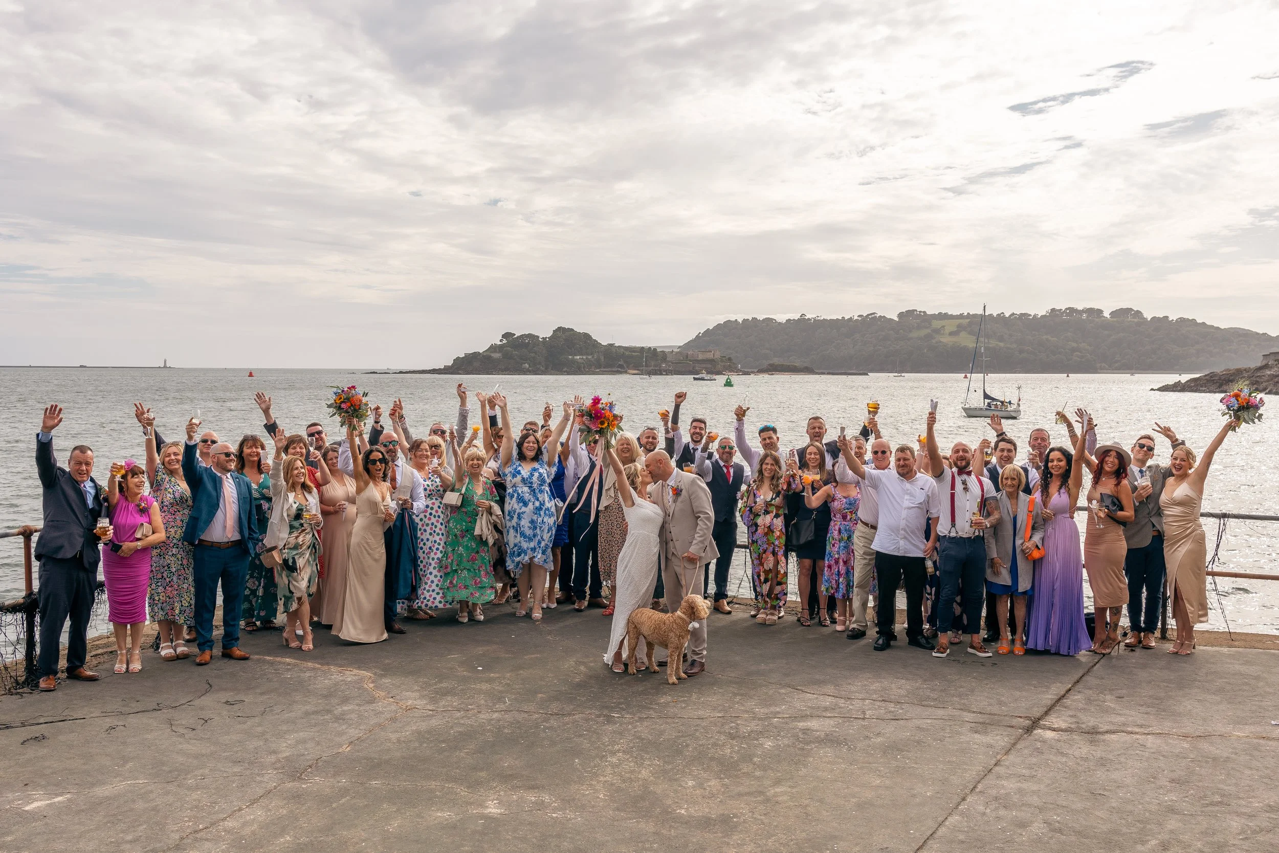 A large group of people celebrating a wedding by the water, with the couple in the center, the bride in a white dress and the groom in a tan suit, surrounded by family and friends, some holding bouquets and glasses, with sailboats and an island in th